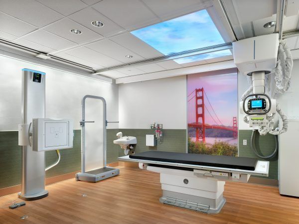 A modern radiology room with an X-ray table, vertical detector, and ceiling-mounted X-ray tube under a sky-themed ceiling.