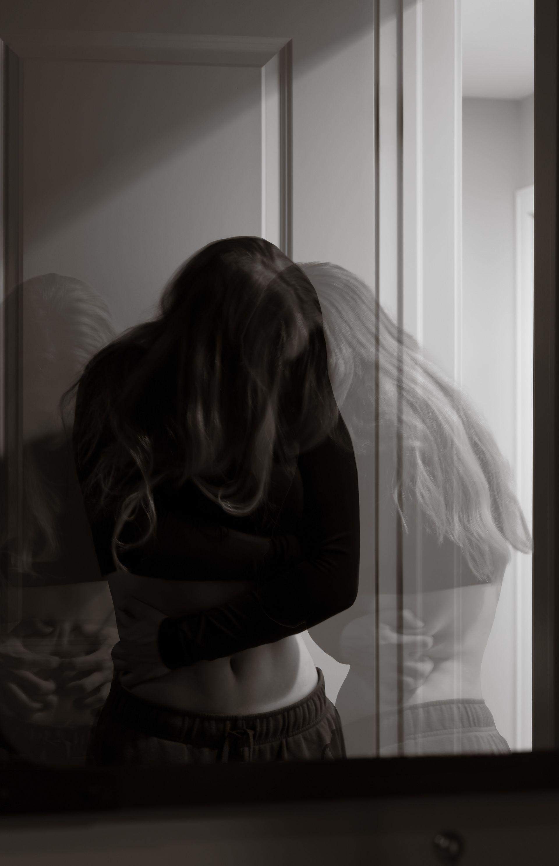A black and white photo of a woman hugging herself in front of a mirror.