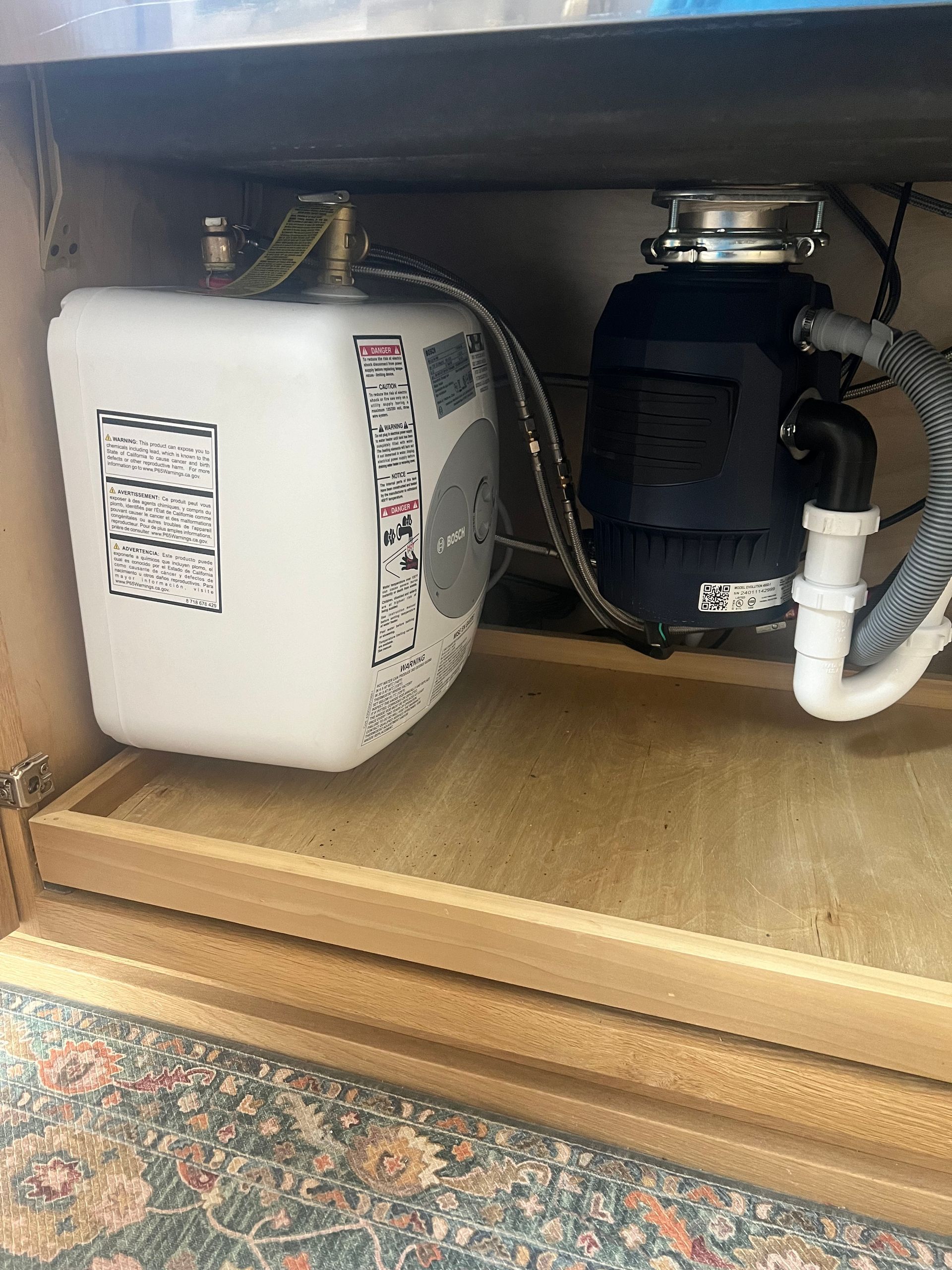 Under-sink view: a small white water heater and black garbage disposal. Plumbing and wiring visible inside a cabinet.