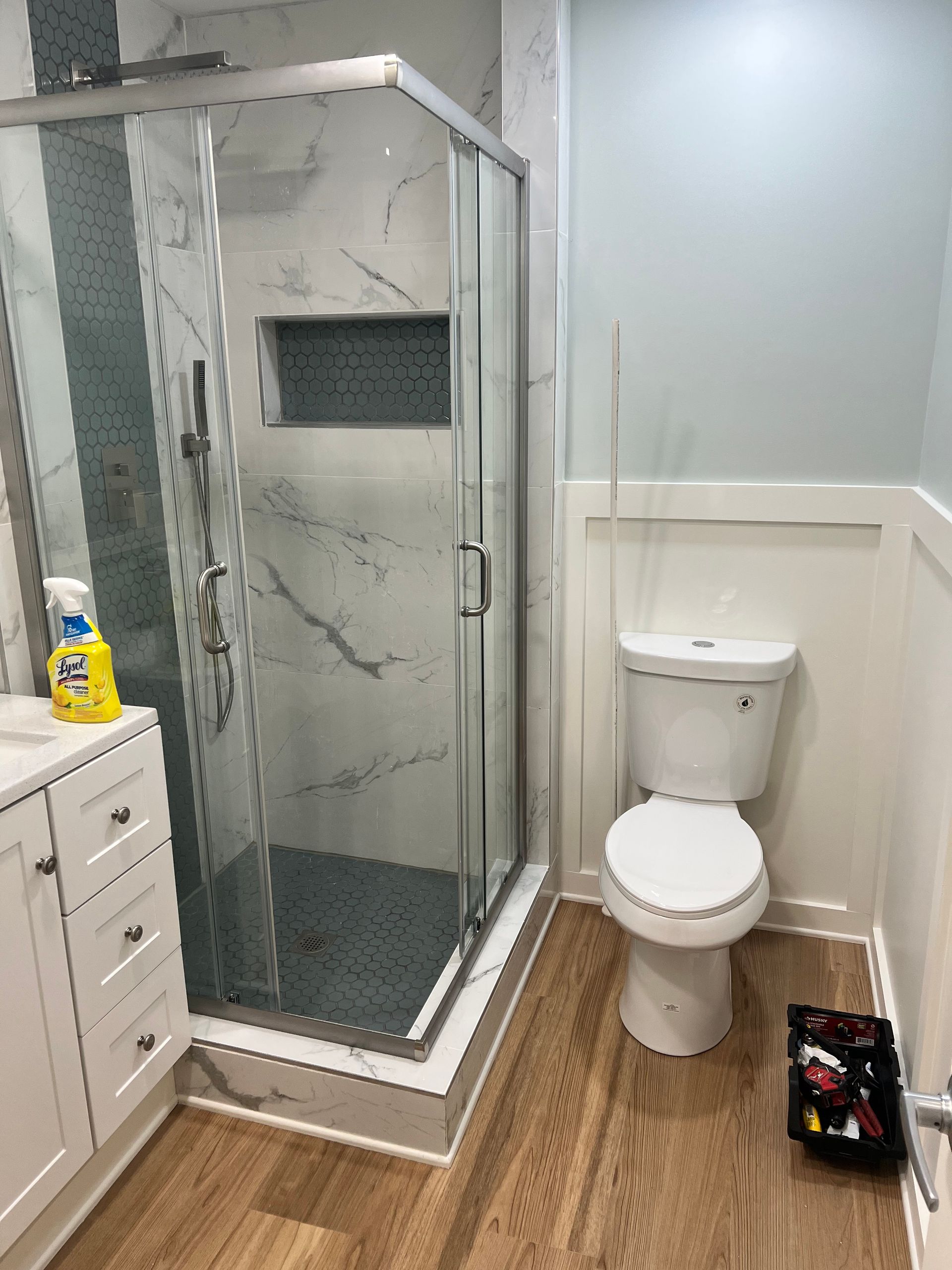 Bathroom with a marble-tiled shower, toilet, and white vanity. Light blue walls and wood-look flooring.
