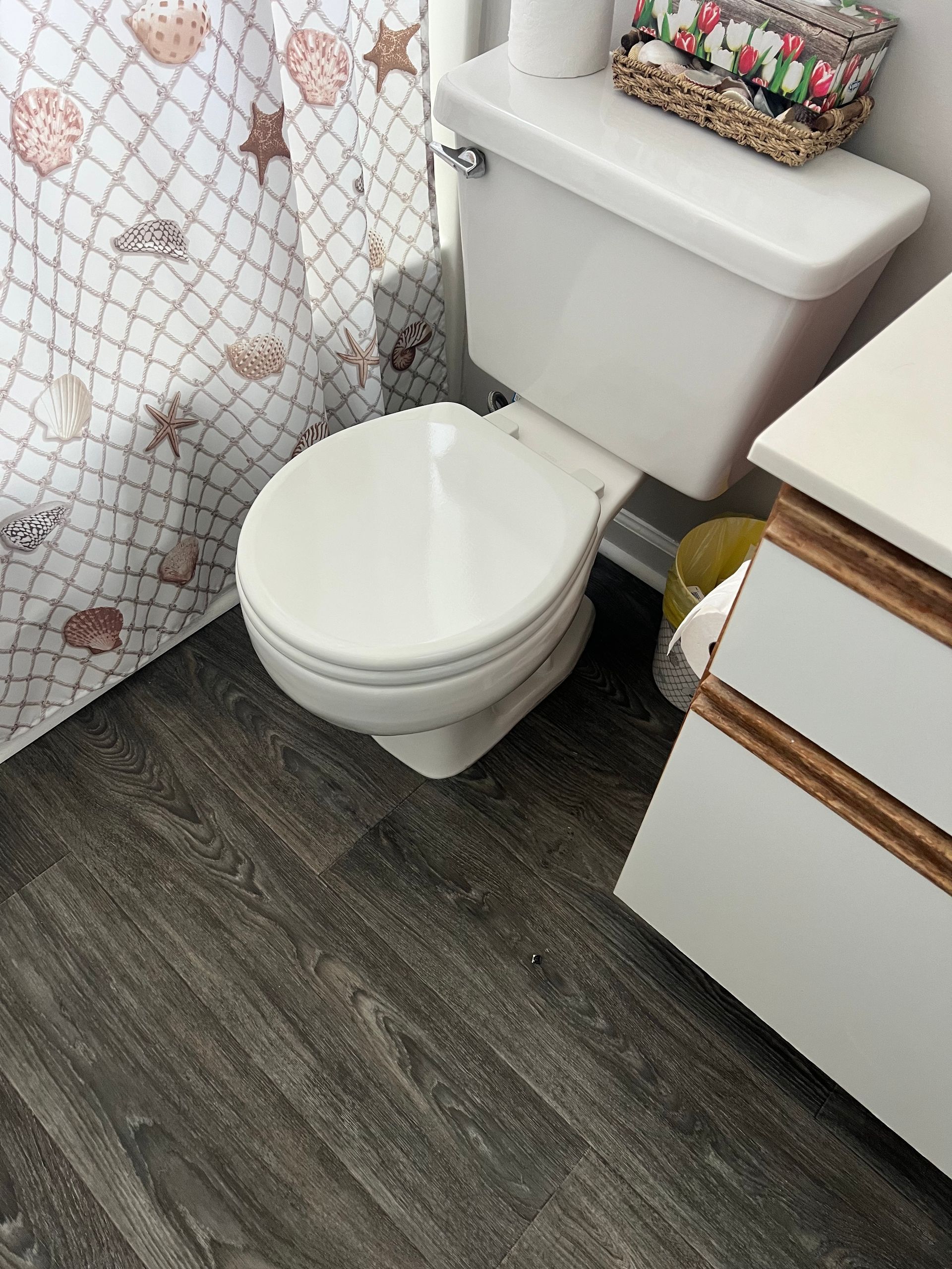 Bathroom with white toilet, gray wood-look floor, and seashell shower curtain.