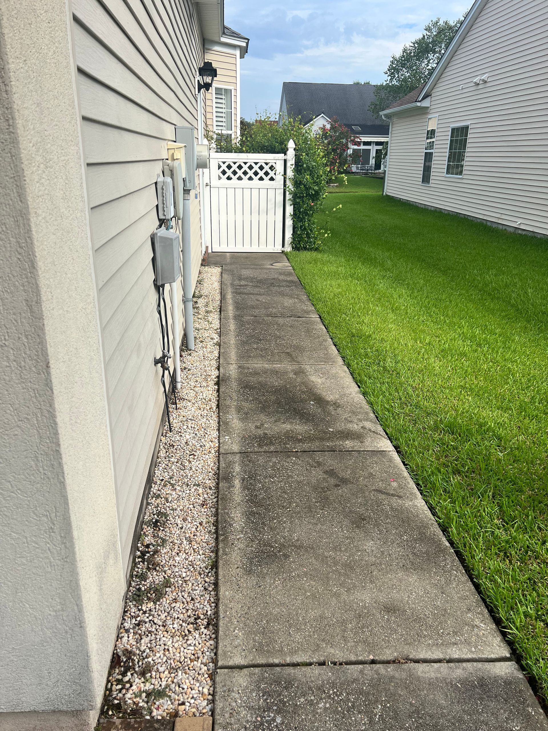 Concrete walkway alongside a house, leading to a white gate and lawn. Gray siding and gravel border.