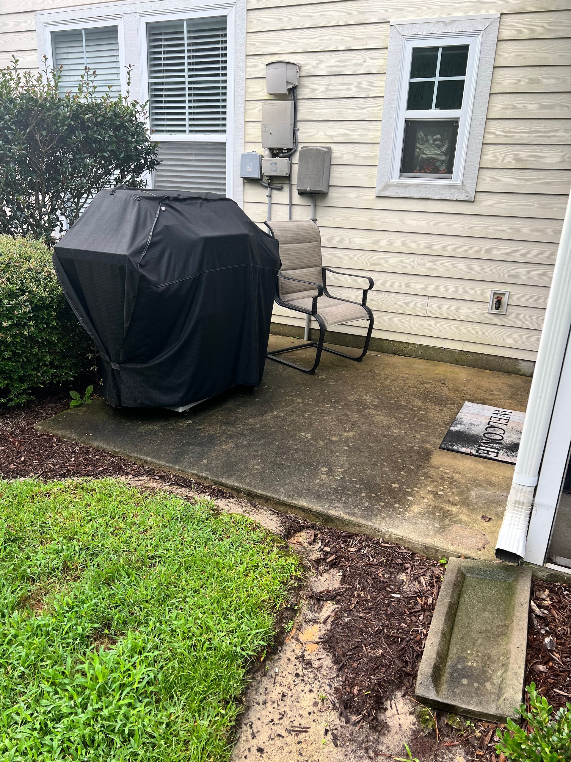 A covered grill and chair sit on a concrete patio next to a cream-colored house.