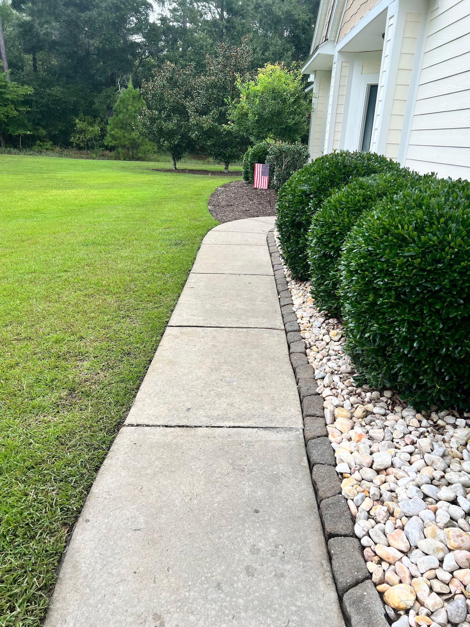 Concrete walkway alongside green lawn and trimmed shrubs, near a building.