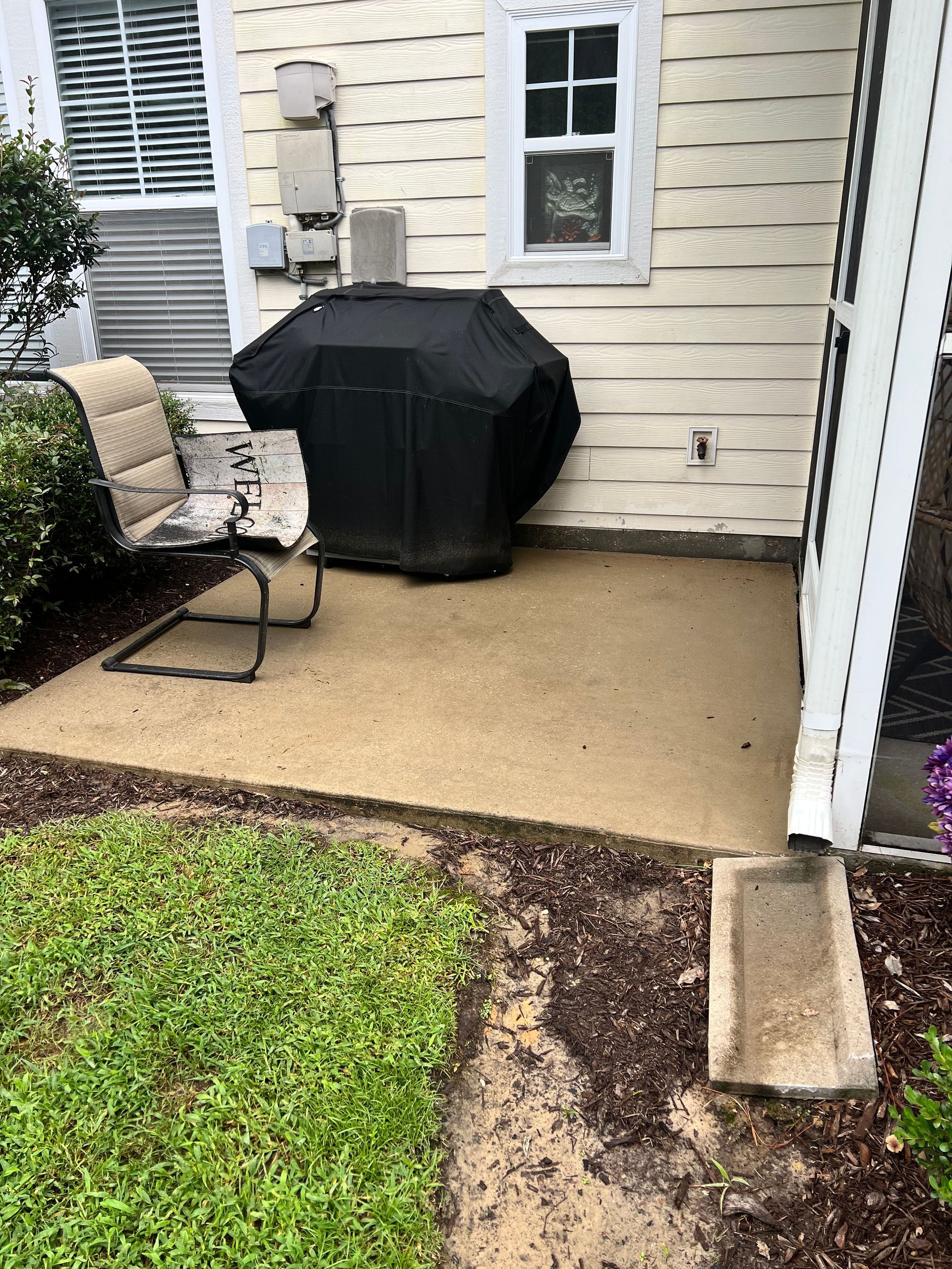 Concrete patio with grill under cover, chair, and adjacent yard.