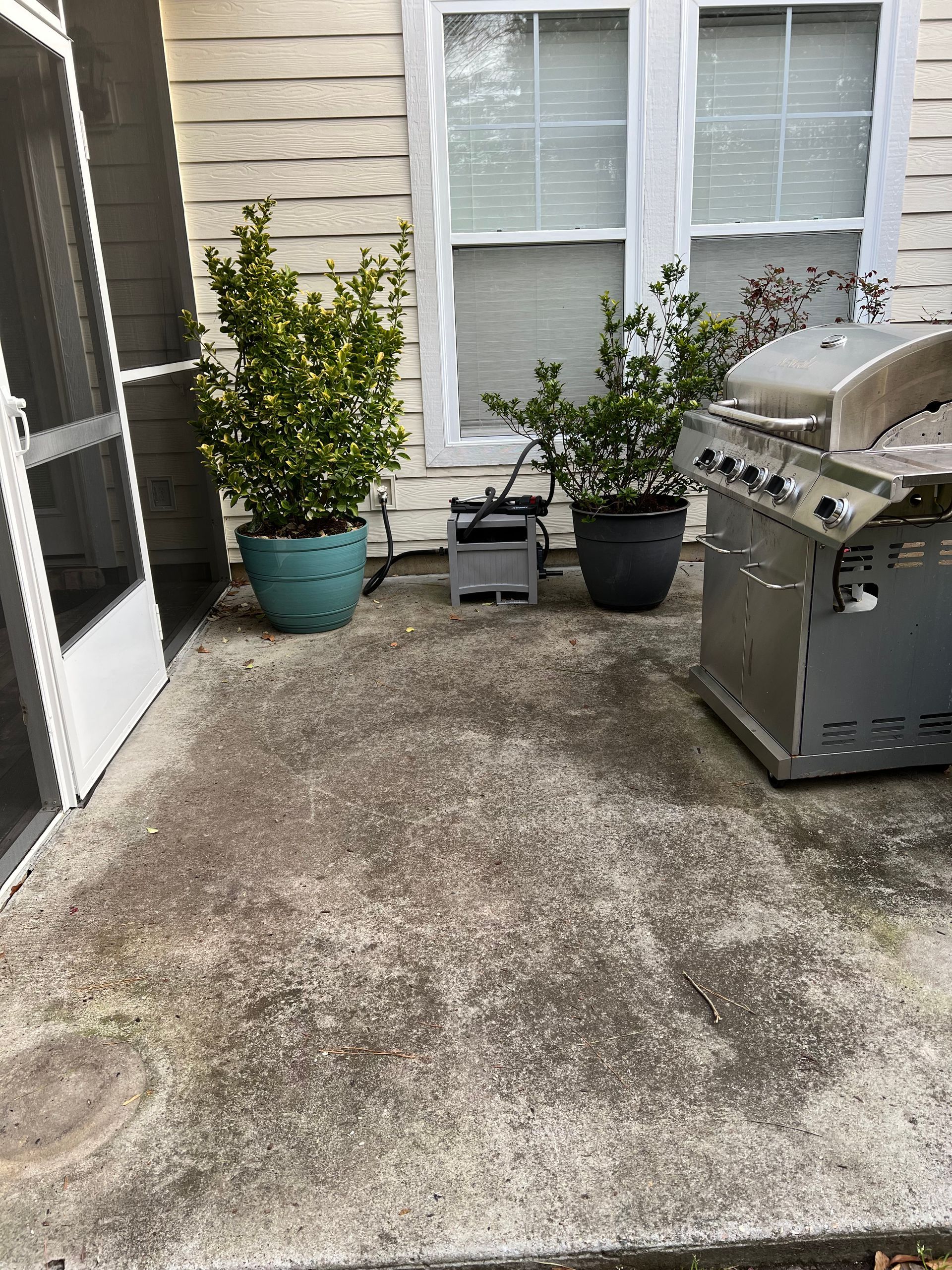 Concrete patio with potted plants, grill, and screen door in front of a white wall with windows.