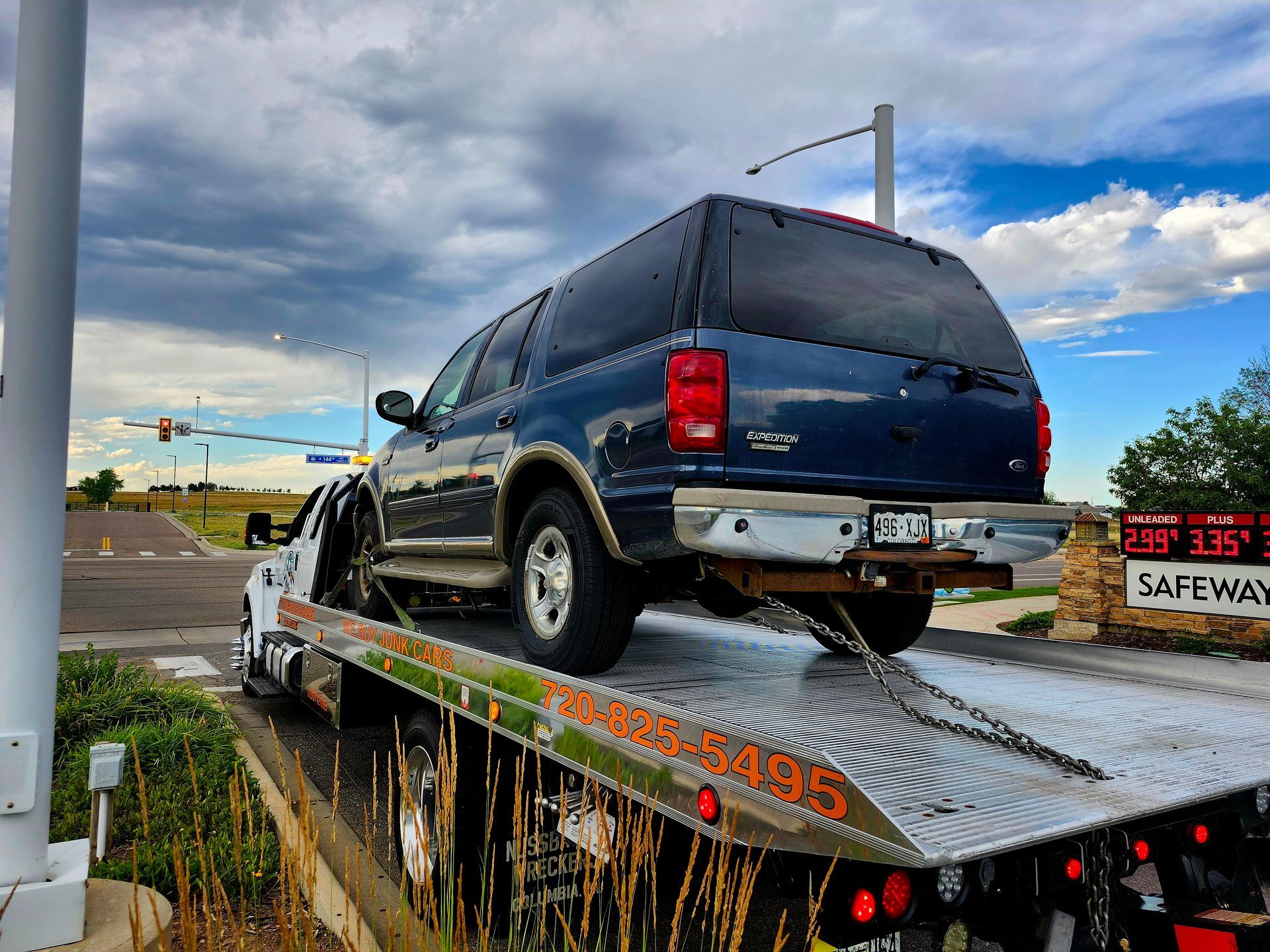 A dark blue SUV is secured onto the flatbed of a tow truck in front of a Safeway gas station under a cloudy sky.