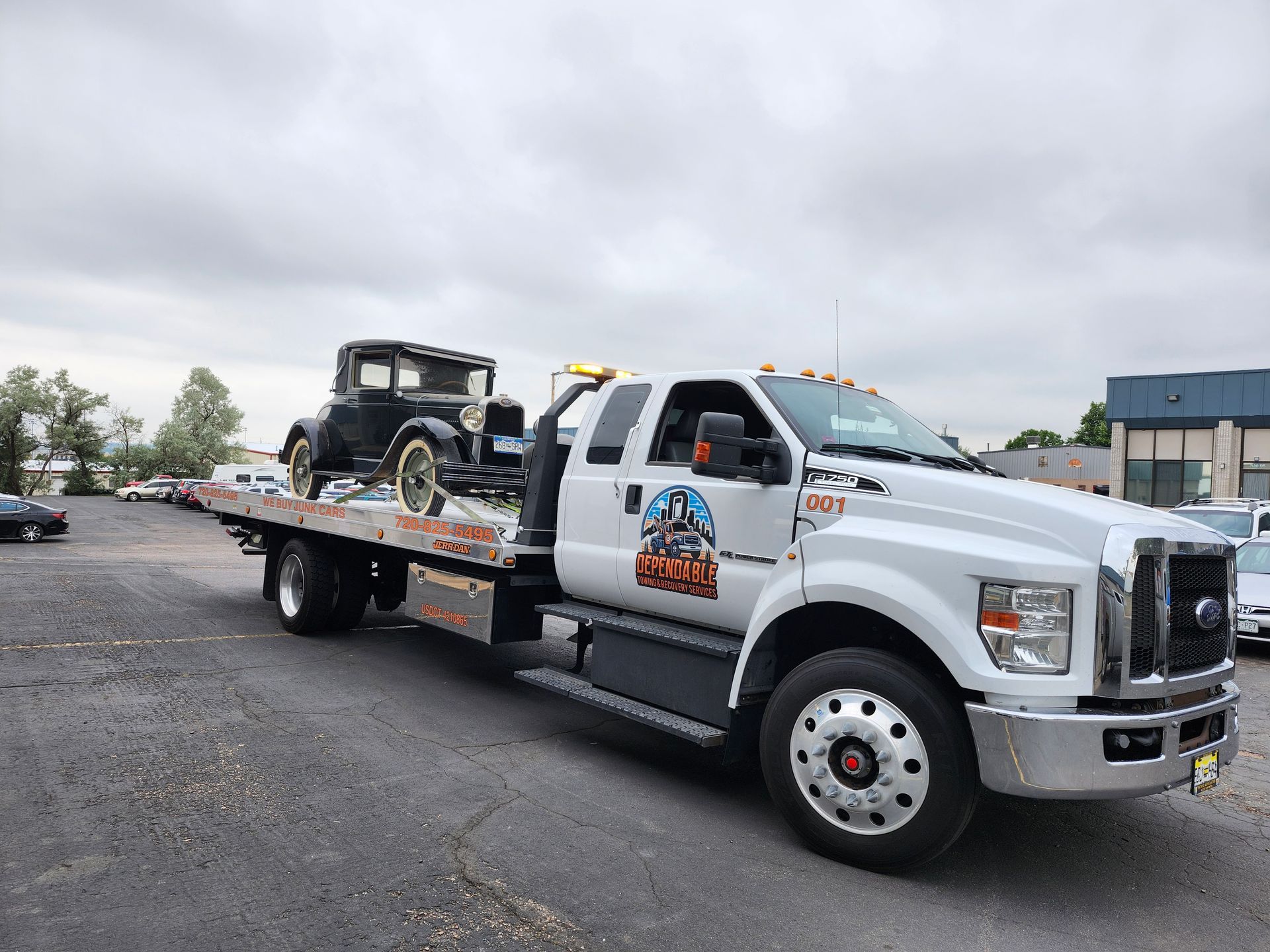 A white flatbed tow truck carries a vintage black coupe across a gravel lot under a cloudy sky.