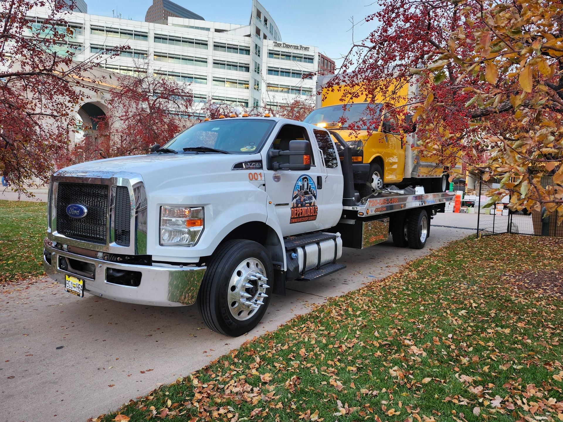 A white flatbed tow truck carrying a yellow delivery van on a path lined with autumn trees and a large building nearby.