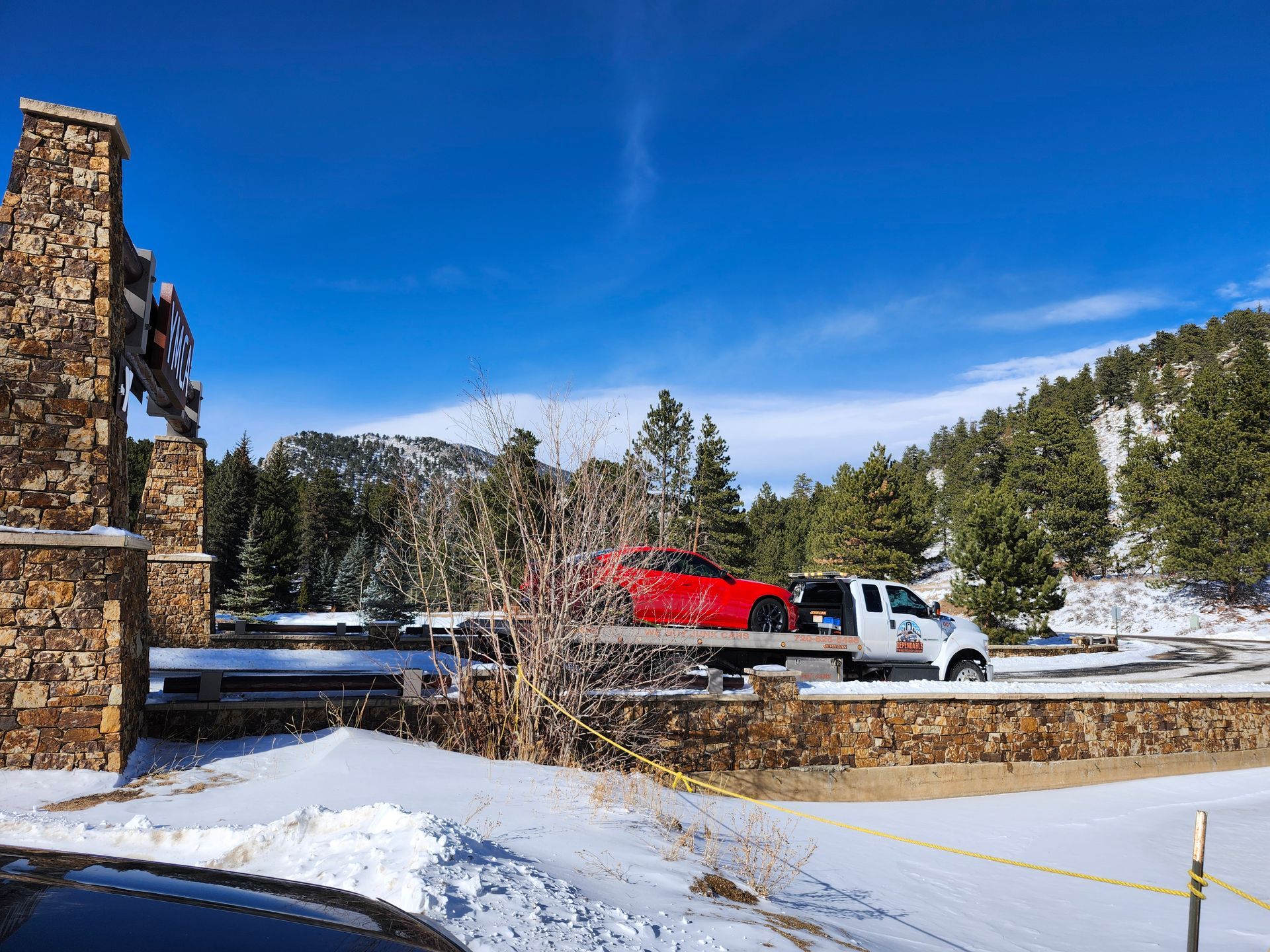 A red sports car sits on a flatbed tow truck in a snow-covered mountain area with stone pillars in the foreground.