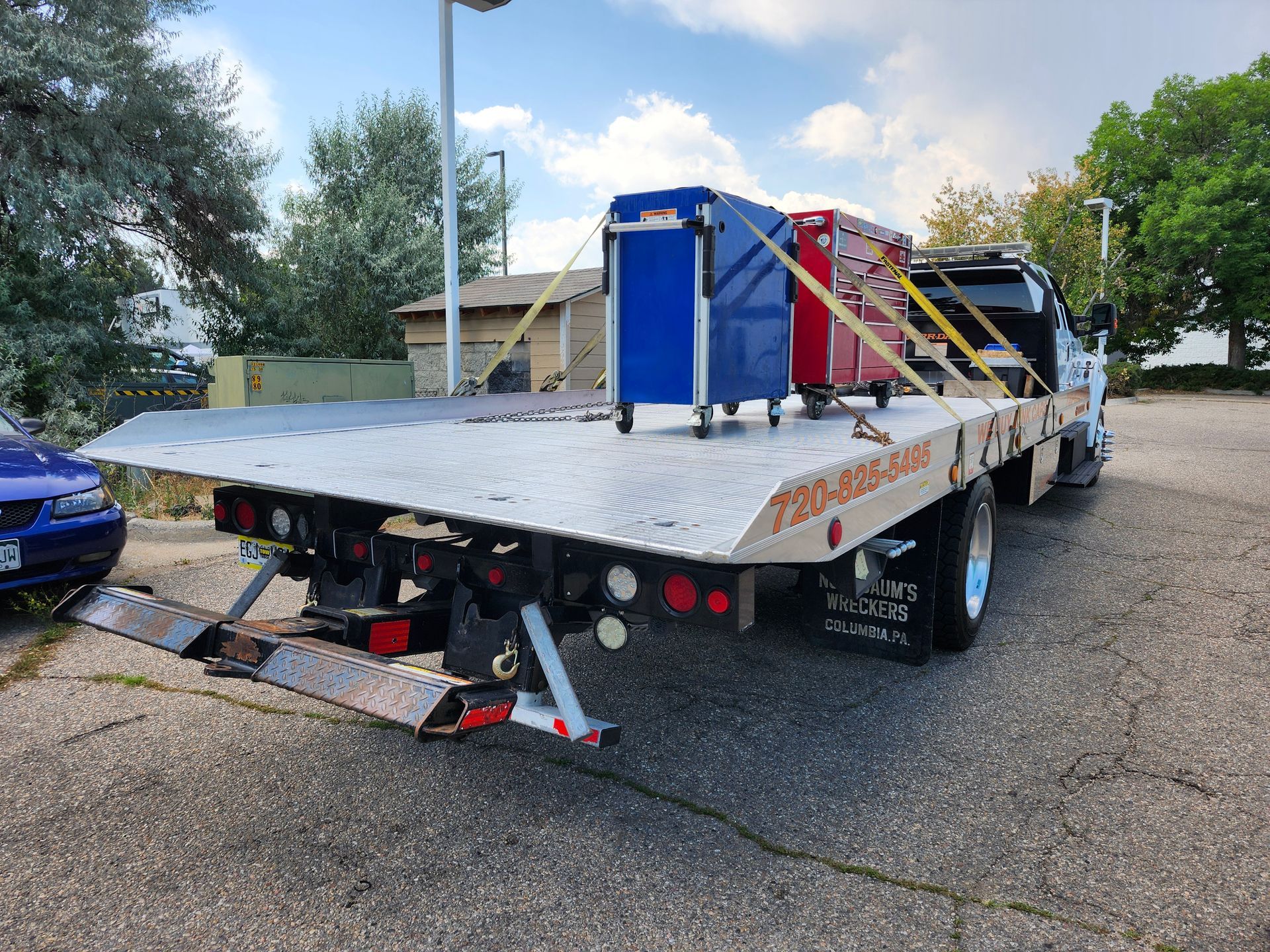 A flatbed tow truck parked on an asphalt lot, carrying a blue rolling tool chest and a smaller red toolbox secured by straps.