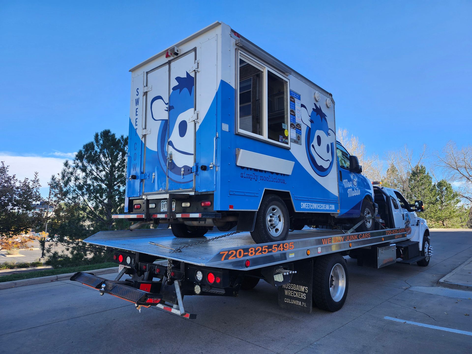 A blue and white food truck with a cartoon logo is being transported on the flatbed of a white tow truck.