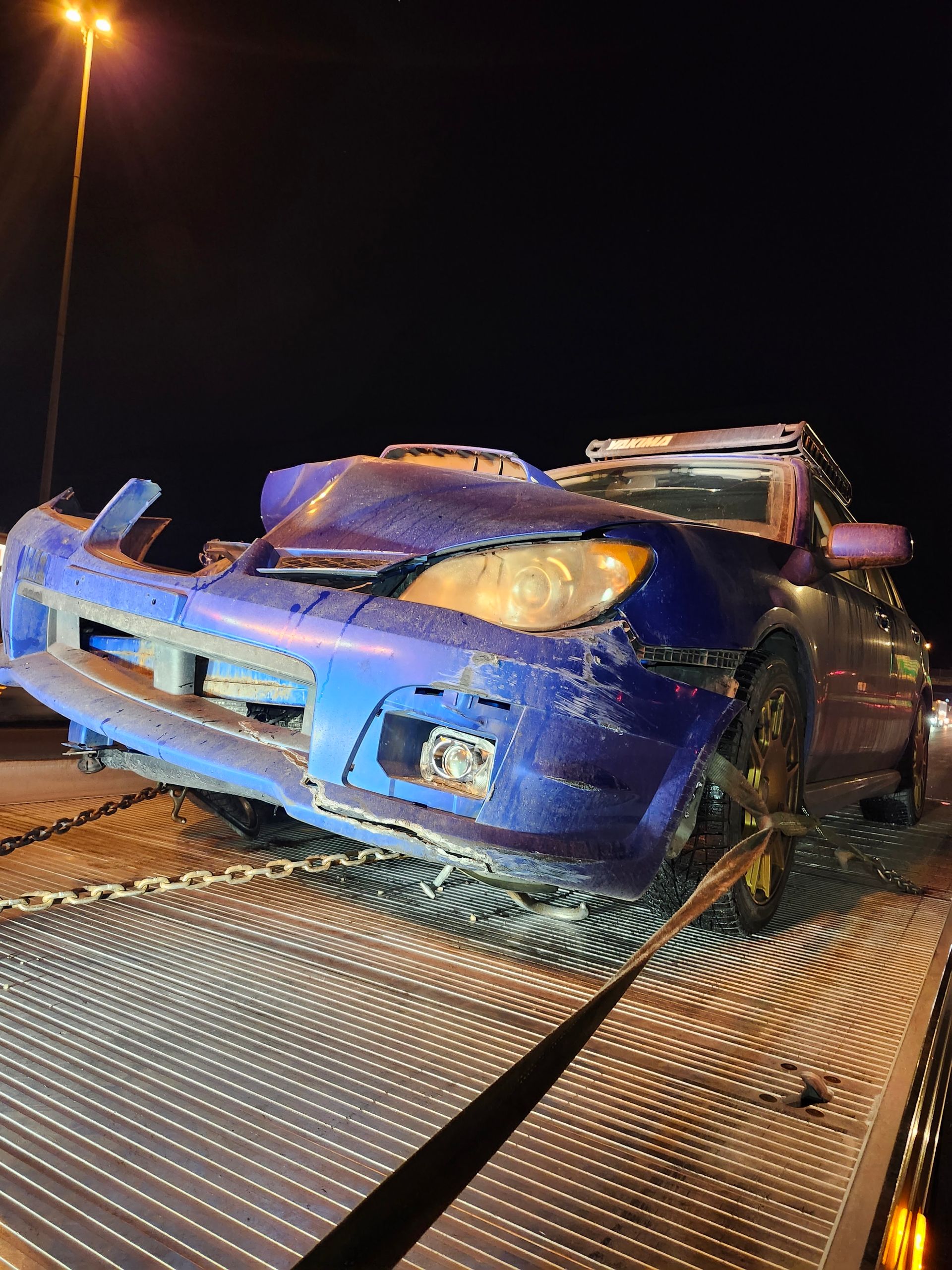 A damaged blue Subaru sedan is secured on the bed of a tow truck at night.