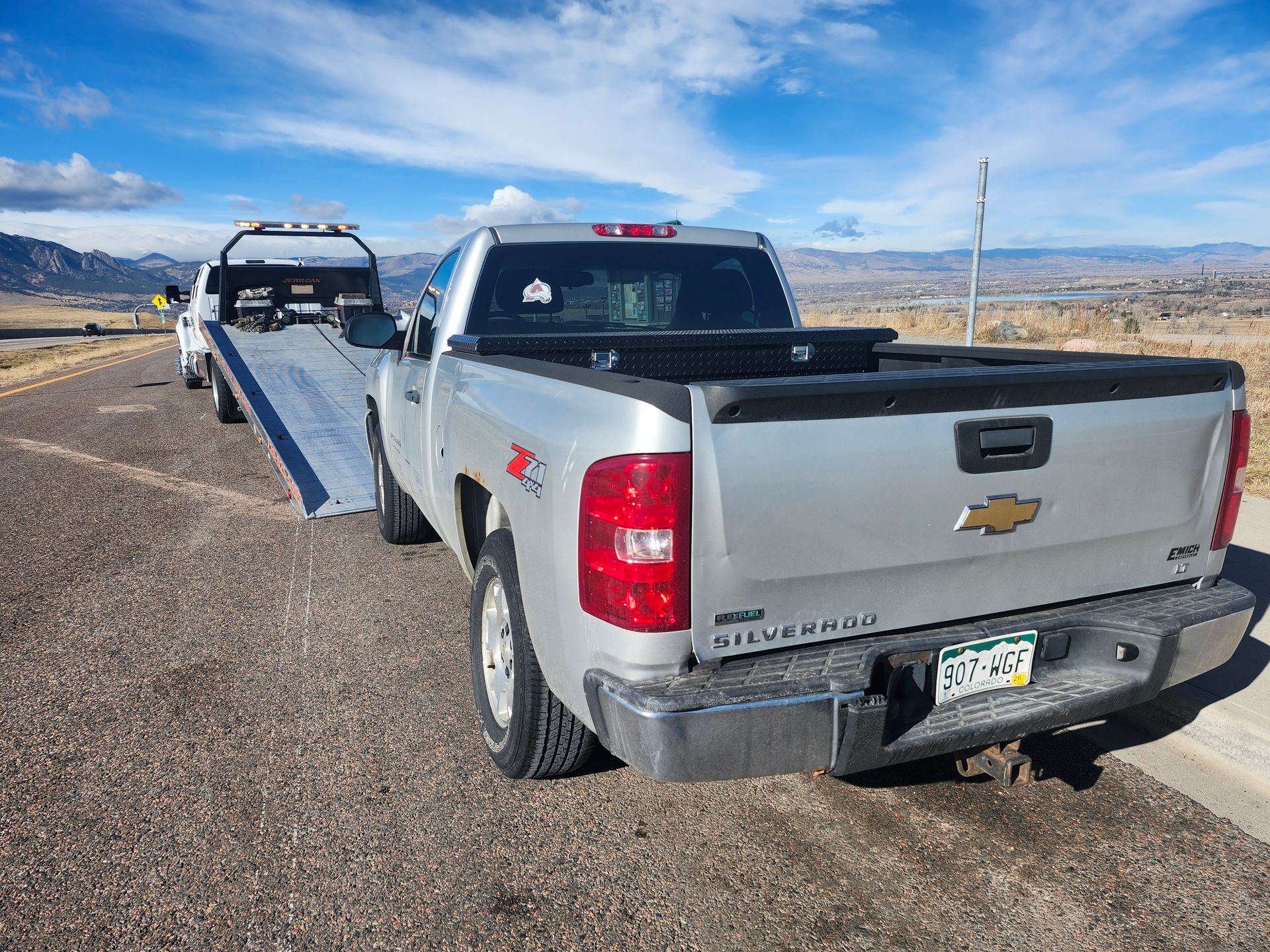 A silver Chevrolet Silverado pickup truck is parked on a dirt shoulder behind a flatbed tow truck with its ramp lowered.