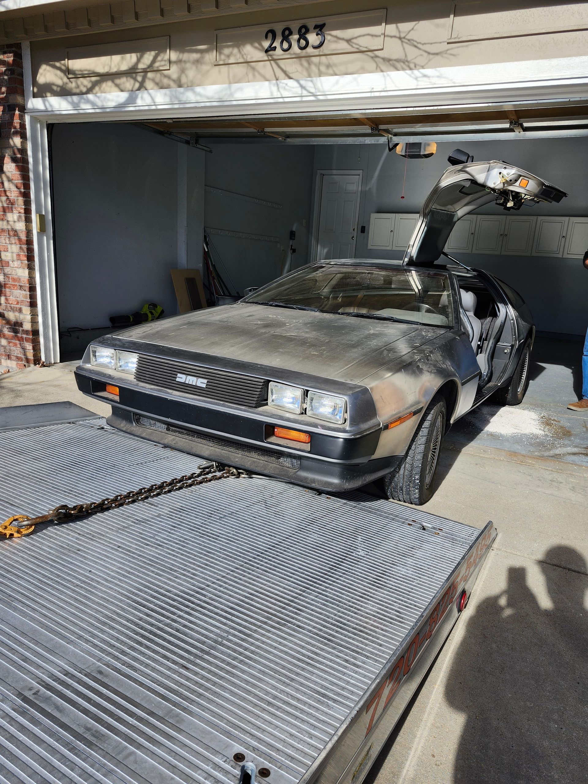 A weathered DeLorean DMC-12 sports car with an open gull-wing door parked inside a garage, facing a flatbed trailer.