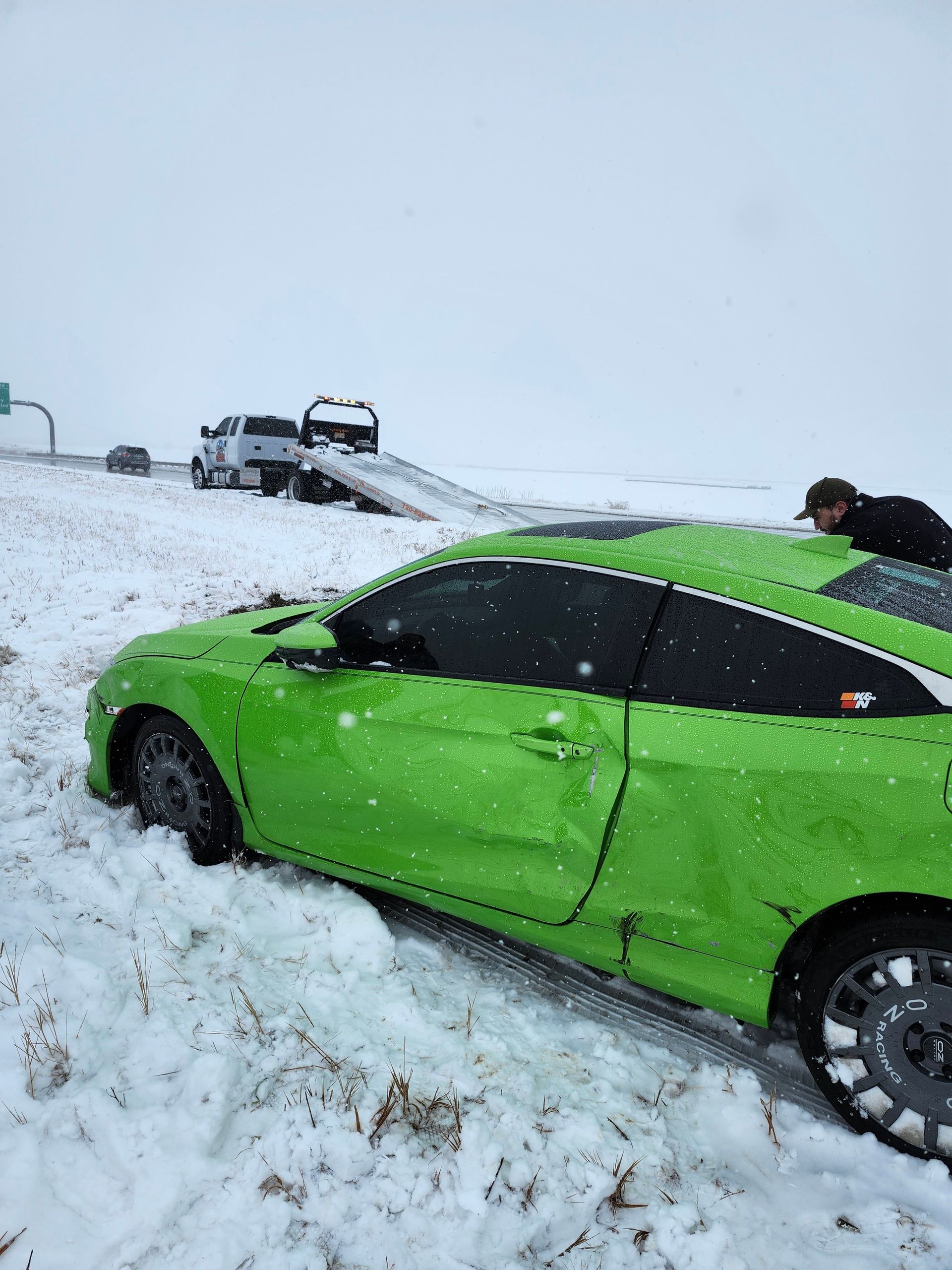 A bright green car sits stuck in deep snow on a roadside, with a tow truck and a person nearby.