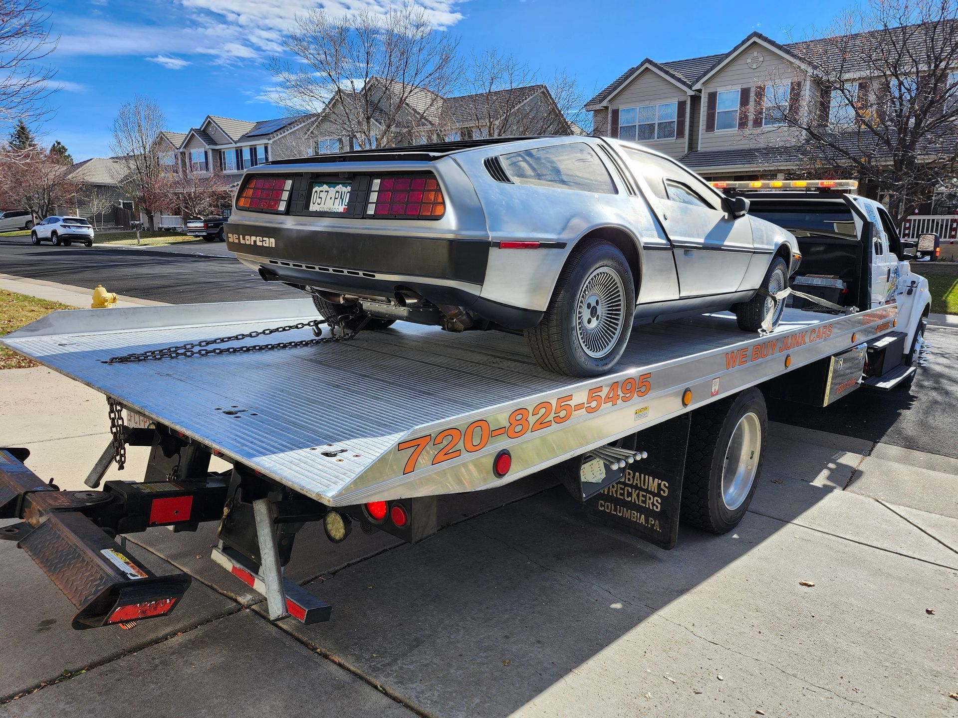A silver DeLorean car sits on the back of a flatbed tow truck parked on a residential street.