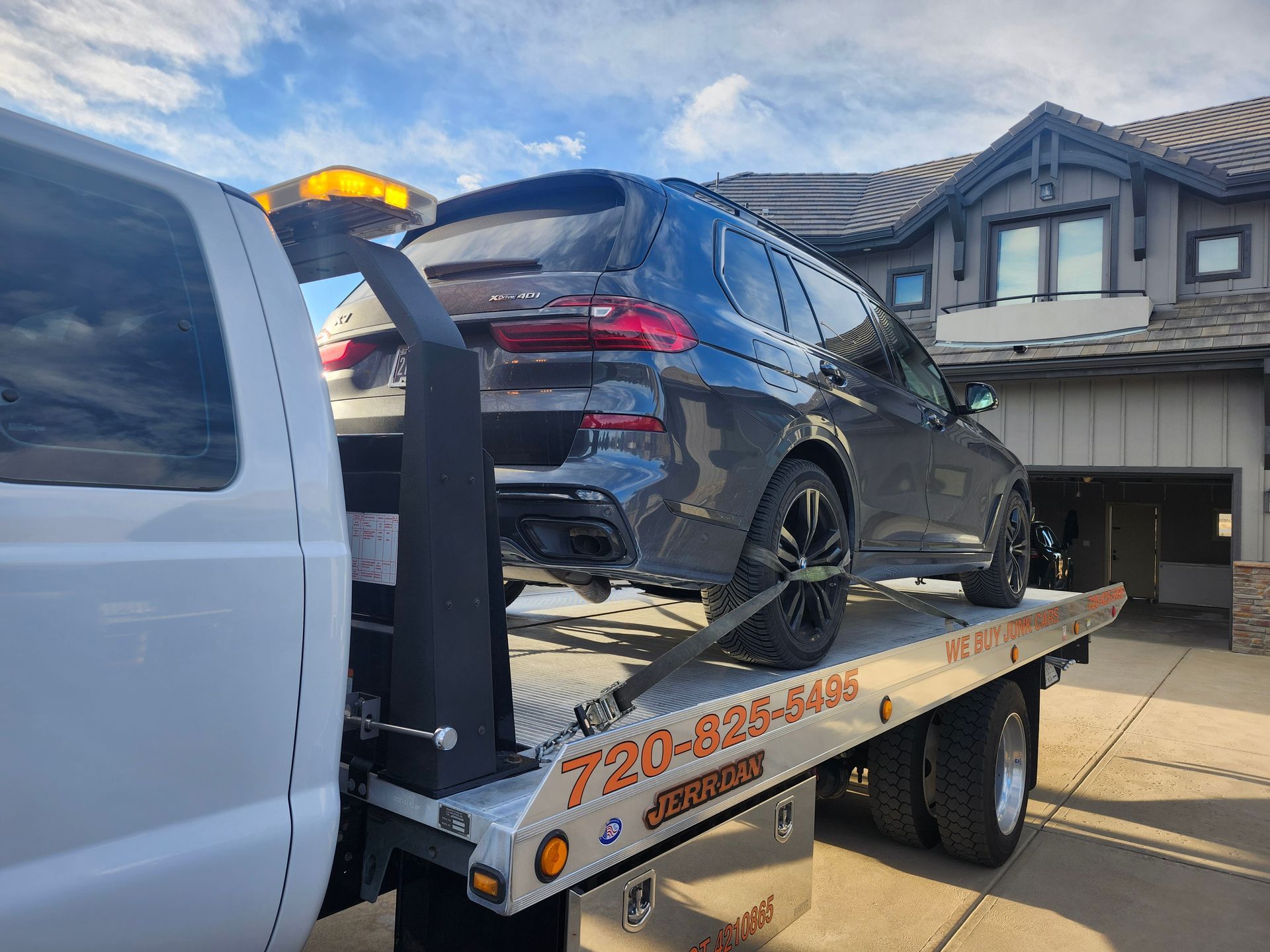 A dark gray BMW SUV loaded onto a white flatbed tow truck in front of a residential home.