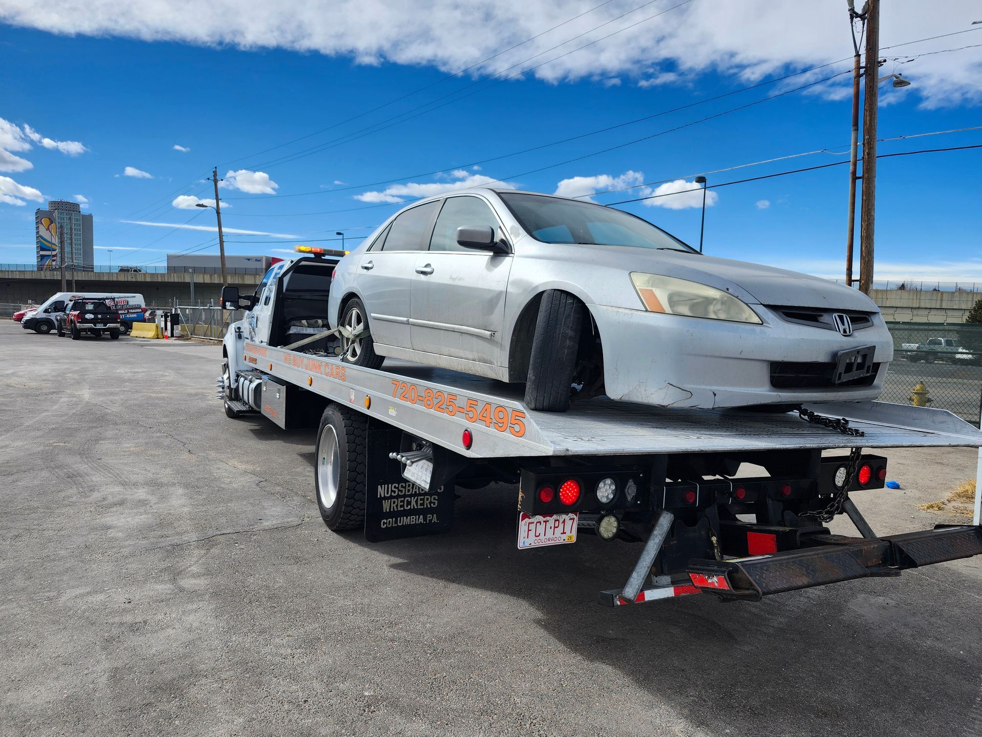 A silver sedan with a flat, angled front tire loaded onto a flatbed tow truck parked on a gravel lot under a sunny sky.
