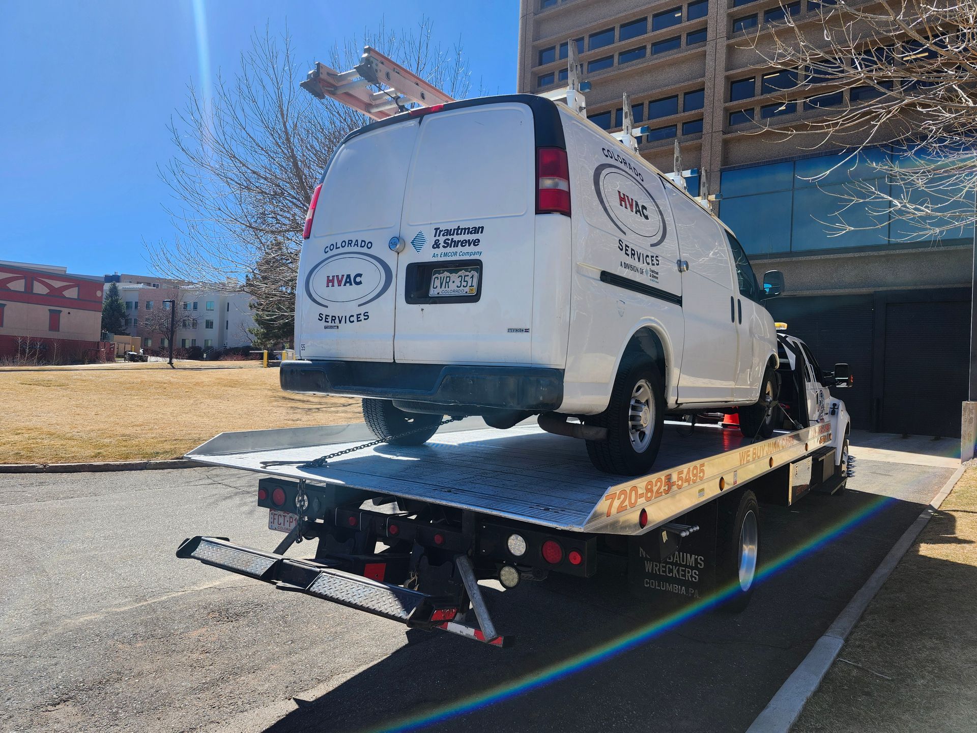 A white work van with logo decals loaded onto a flatbed tow truck parked near a multi-story building.
