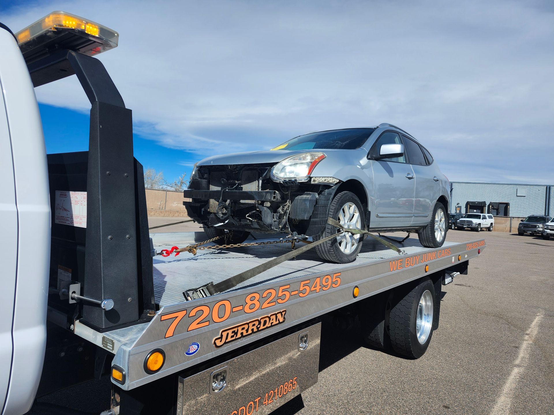 A silver SUV with missing front bumper parts is secured to the bed of a flatbed tow truck on a sunny day.