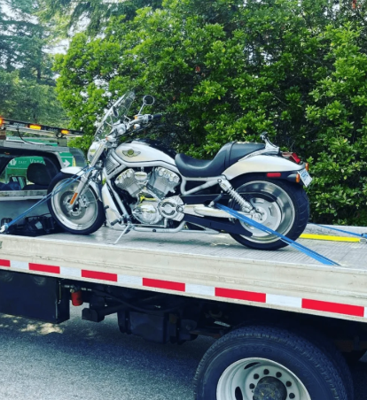 A white Harley-Davidson V-Rod motorcycle secured with blue tie-down straps on the back of a flatbed tow truck.