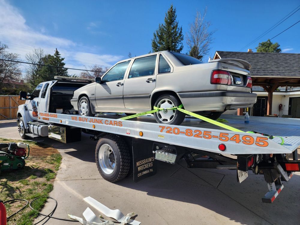 A silver Volvo sedan secured to the bed of a white flatbed tow truck on a suburban driveway.