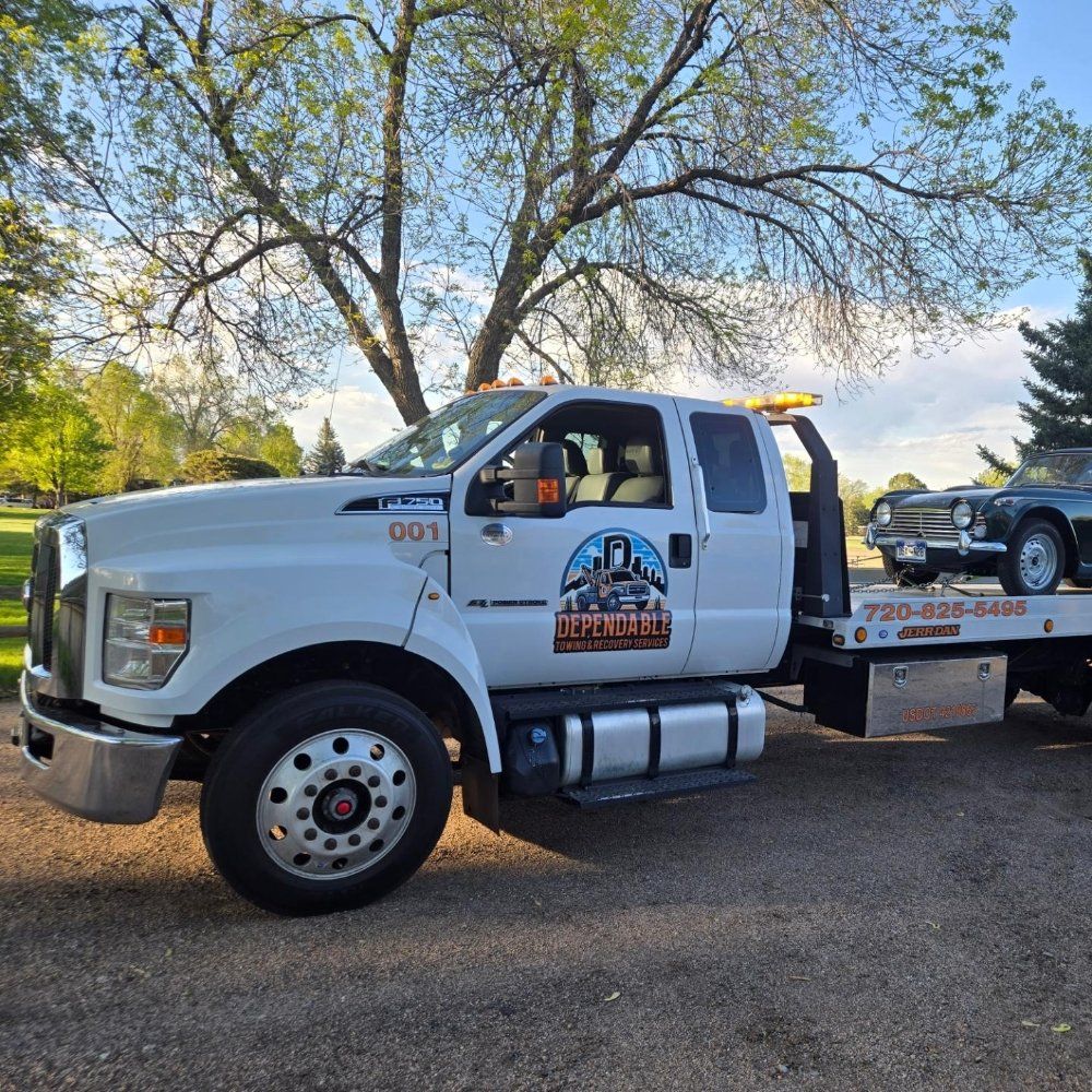 A white Dependable towing truck parked on gravel with a dark classic car secured on its flatbed under sunny trees.