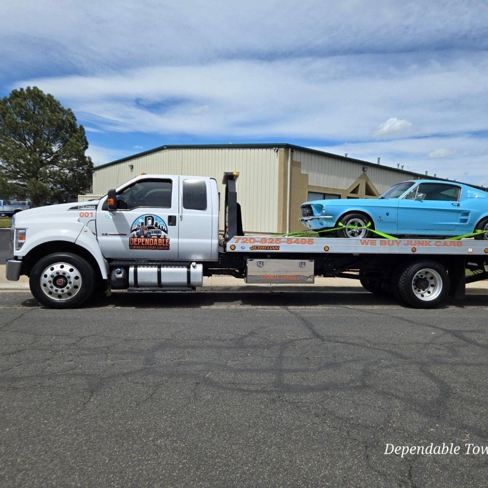 A white tow truck carries a bright blue vintage Ford Mustang on its flatbed in a paved lot under a blue sky.