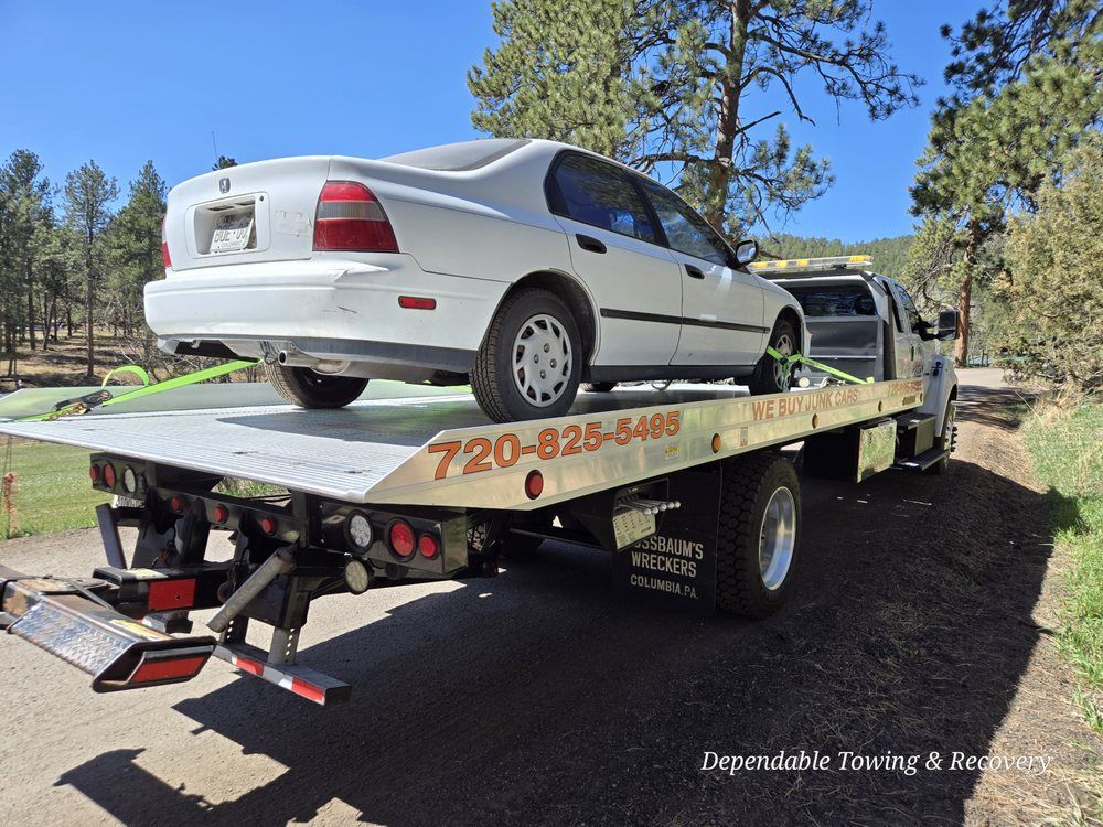 A white sedan loaded onto the back of a flatbed tow truck on a sunny road lined with pine trees.