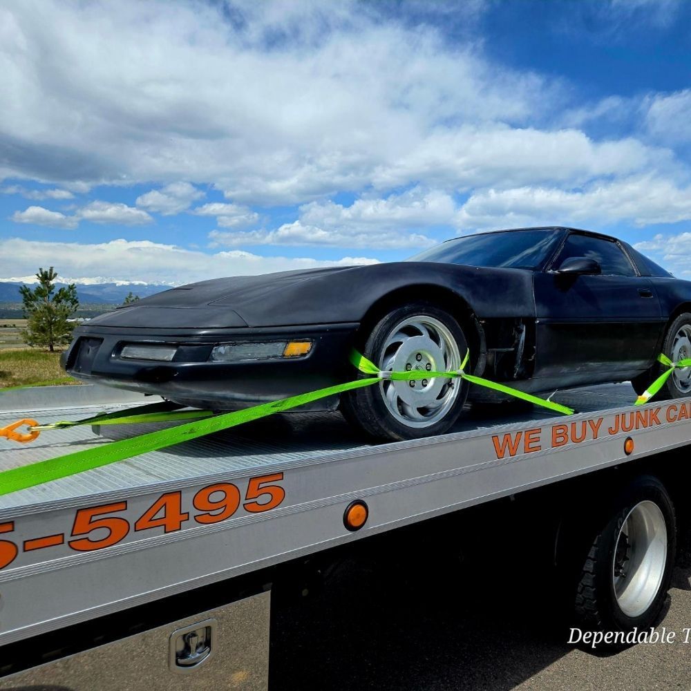 A black Chevrolet Corvette is strapped to a flatbed tow truck against a backdrop of blue skies and distant mountains.