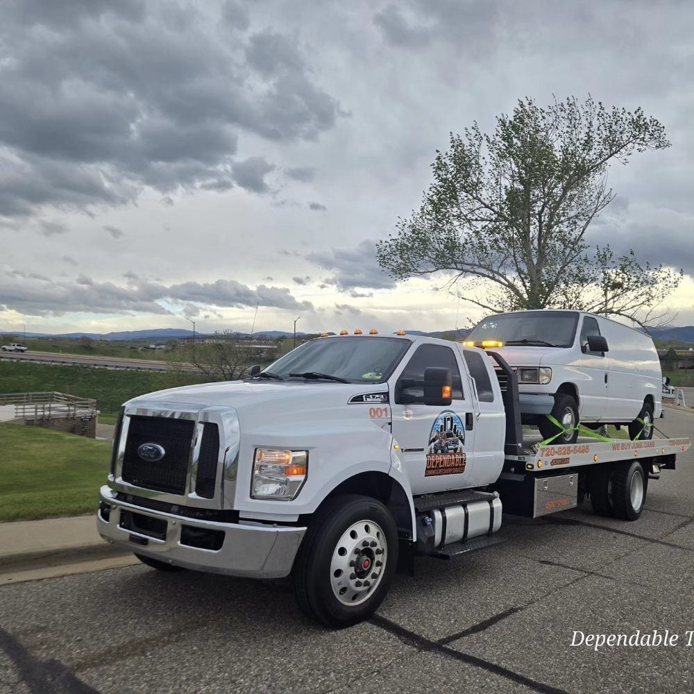 A white Ford flatbed tow truck carrying a white van on its bed parked outdoors under a cloudy sky.