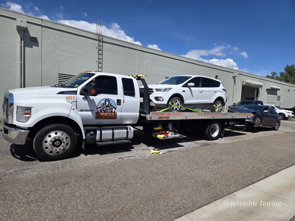 A white flatbed tow truck carrying a white SUV, parked in front of a long, gray industrial building under a blue sky.