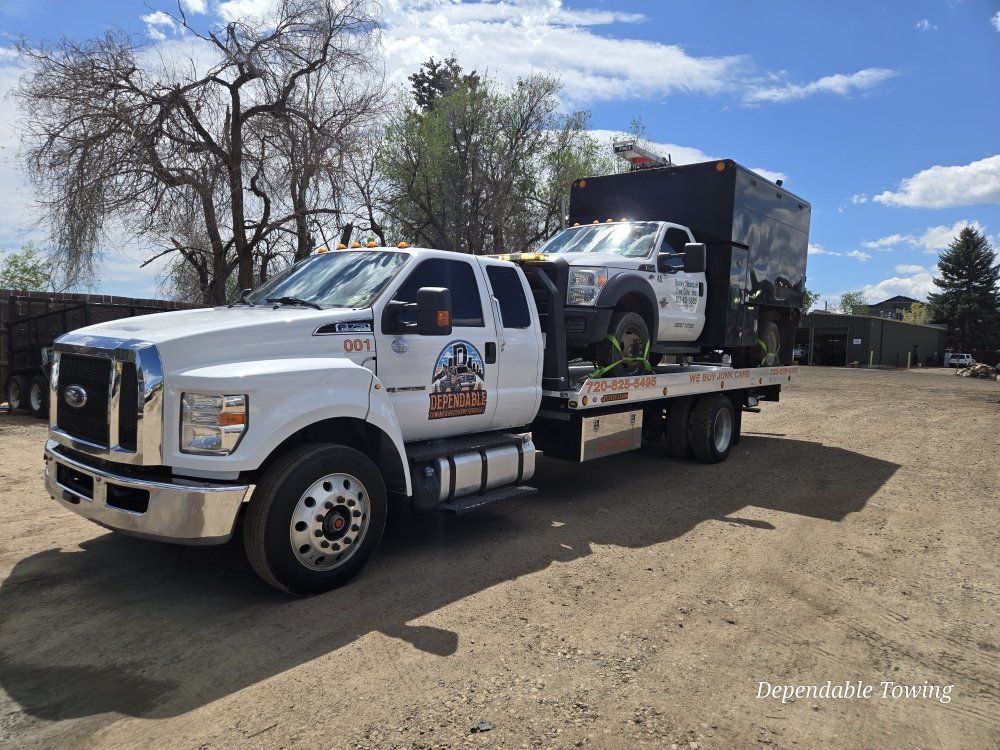 A white flatbed tow truck carrying a white service truck with a large black utility box, parked on a dirt lot.