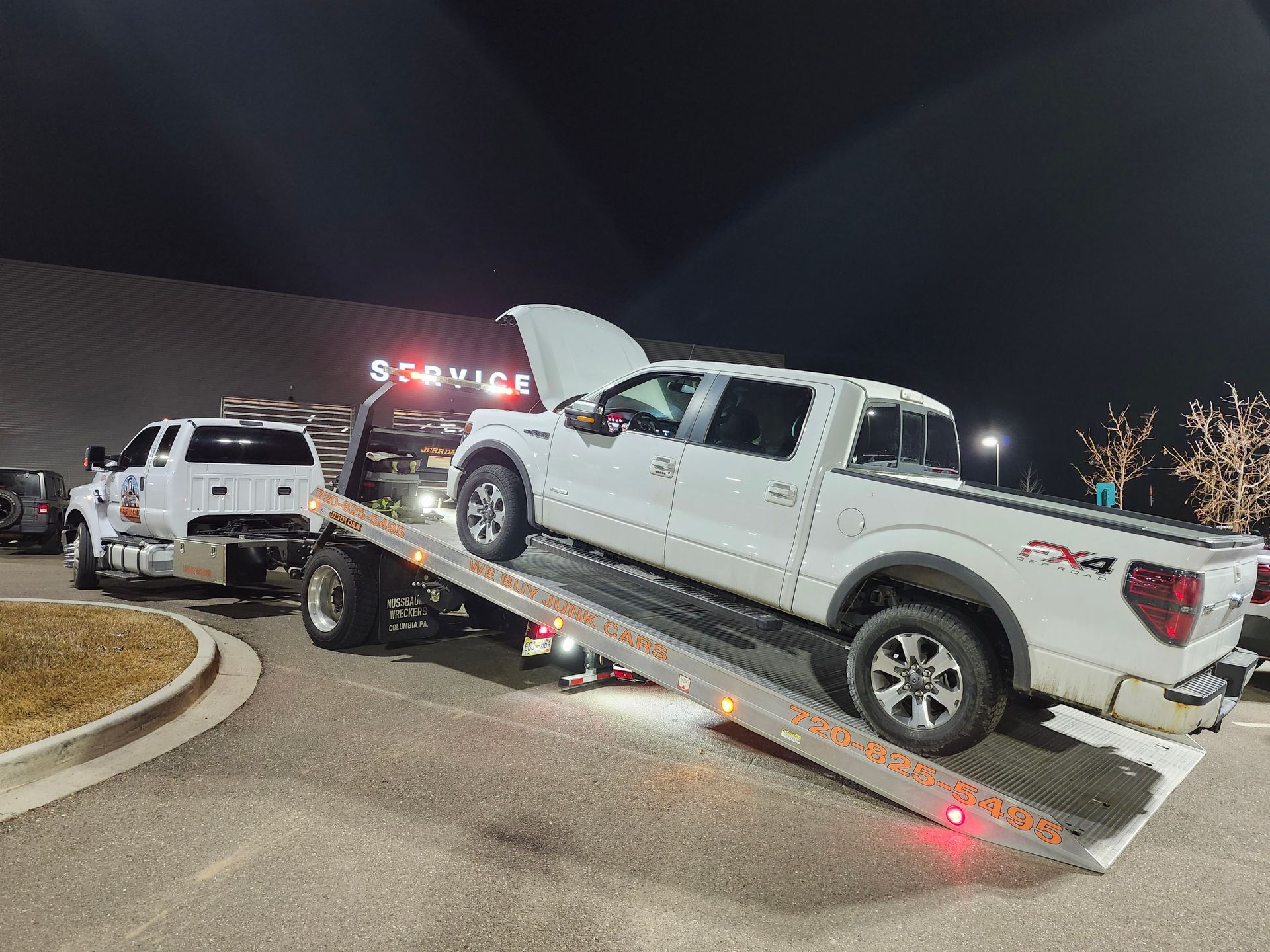 A white pickup truck with its hood open is being winched onto the back of a flatbed tow truck at night.