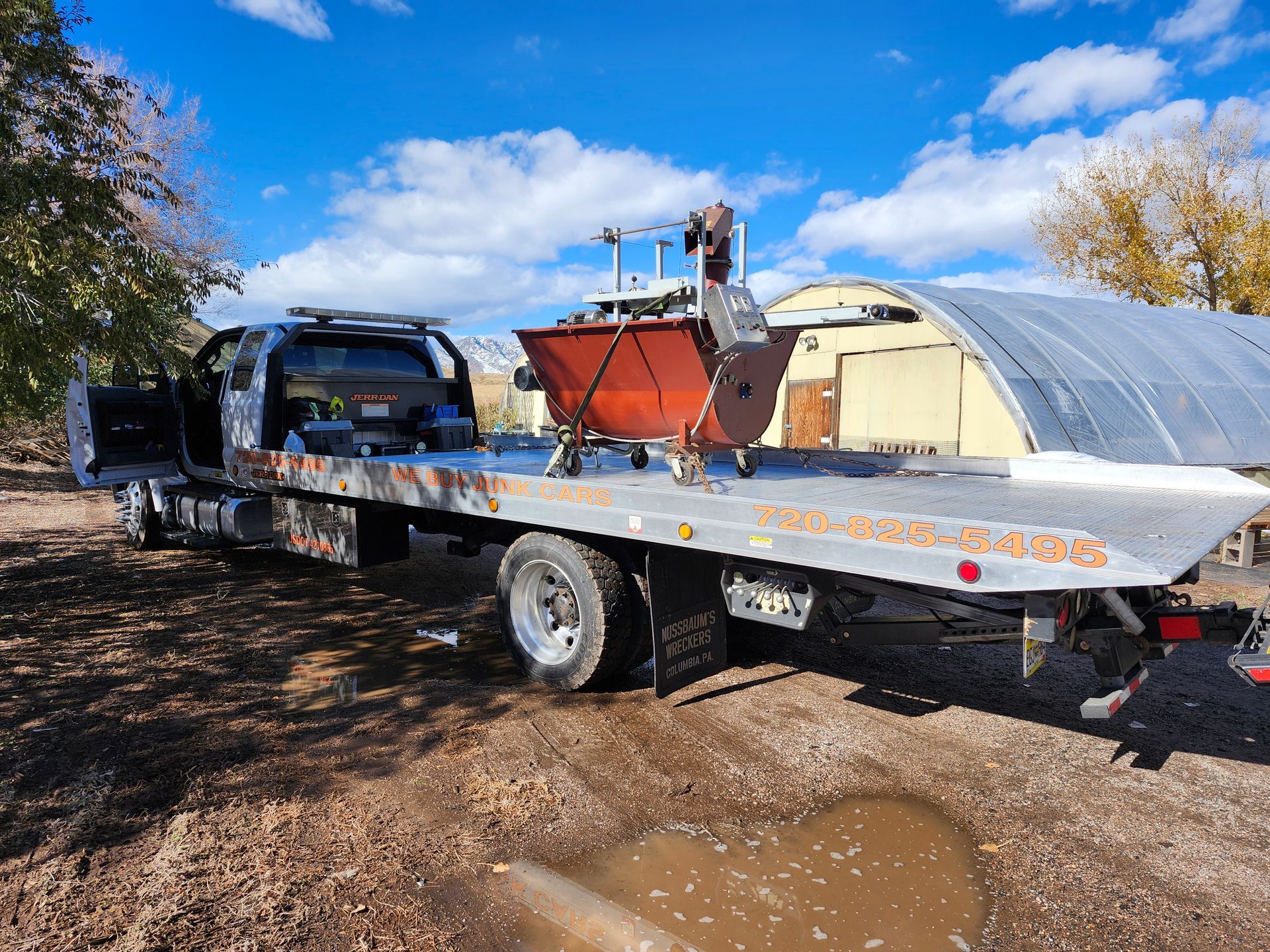 A flatbed tow truck parked outdoors carries a red, wheeled piece of industrial machinery on its deck.