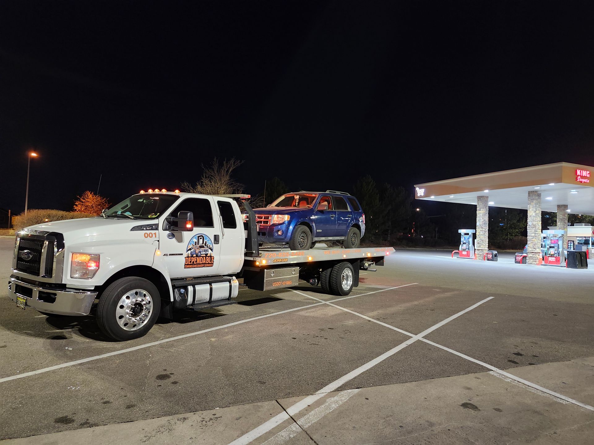 A white tow truck carries a blue SUV at night in front of a brightly lit gas station.