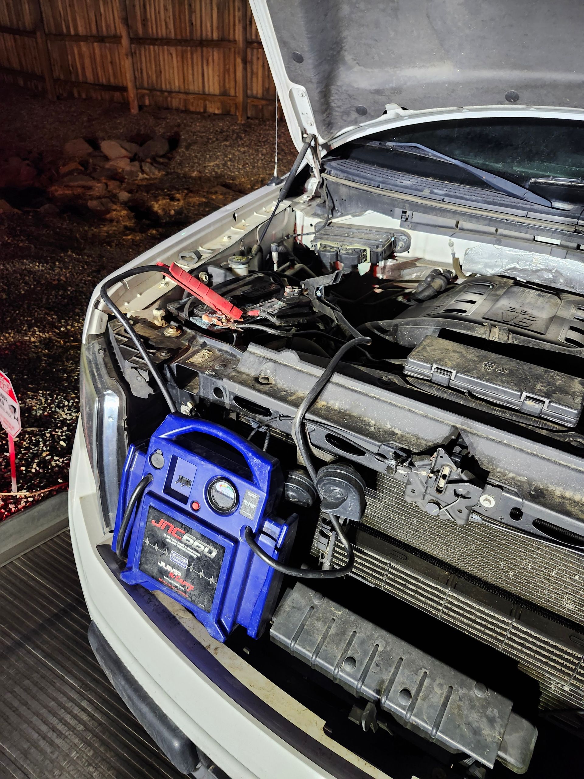 A blue jump starter unit sits on a vehicle's front bumper, with cables connected to the battery under the open hood.