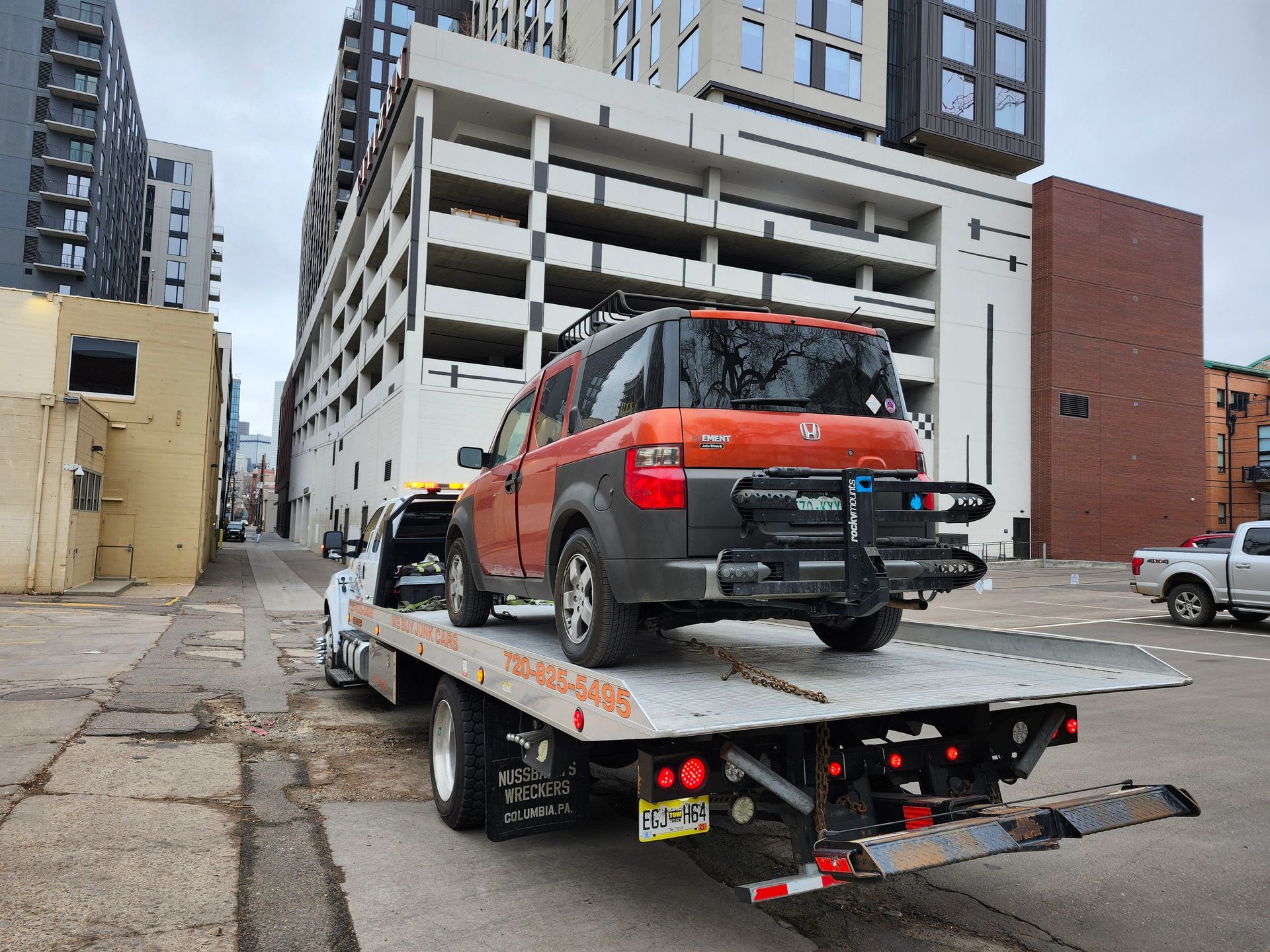 An orange Honda Element with a bike rack on the back is loaded onto a flatbed tow truck parked in an urban alley.