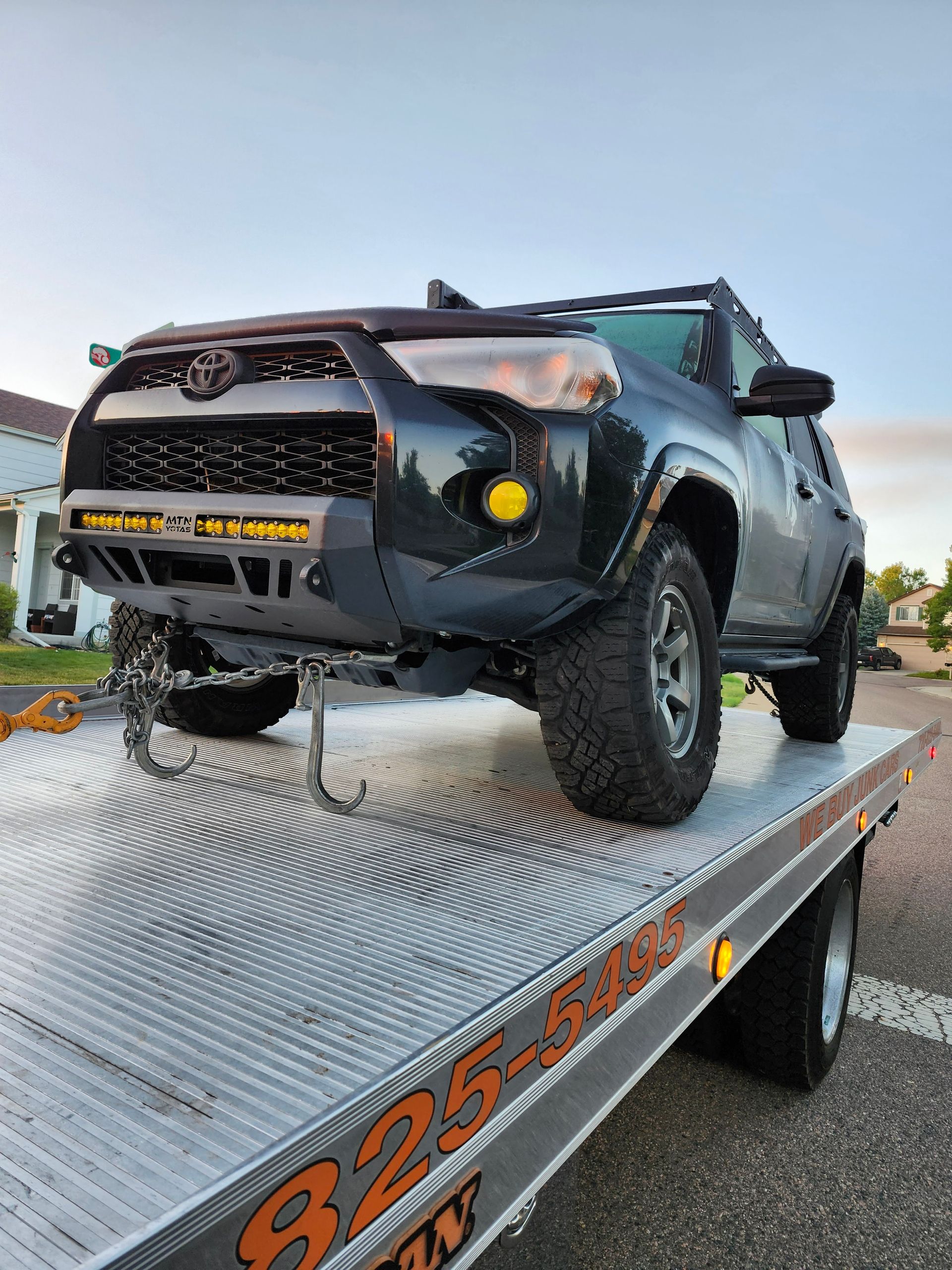 A dark Toyota SUV secured with chains to the bed of a metal flatbed tow truck on a suburban street.