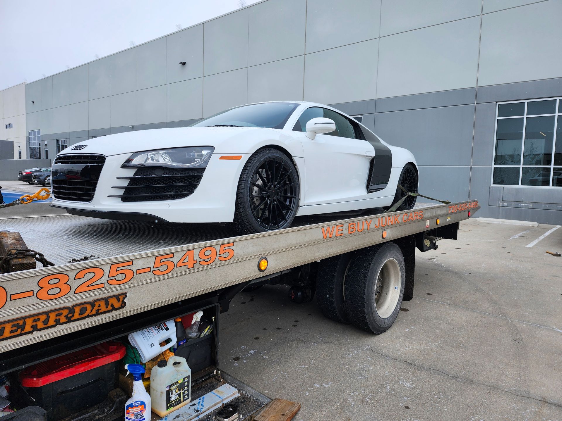 A white Audi R8 sports car with black rims is parked on the flatbed of a tow truck outside a large industrial building.