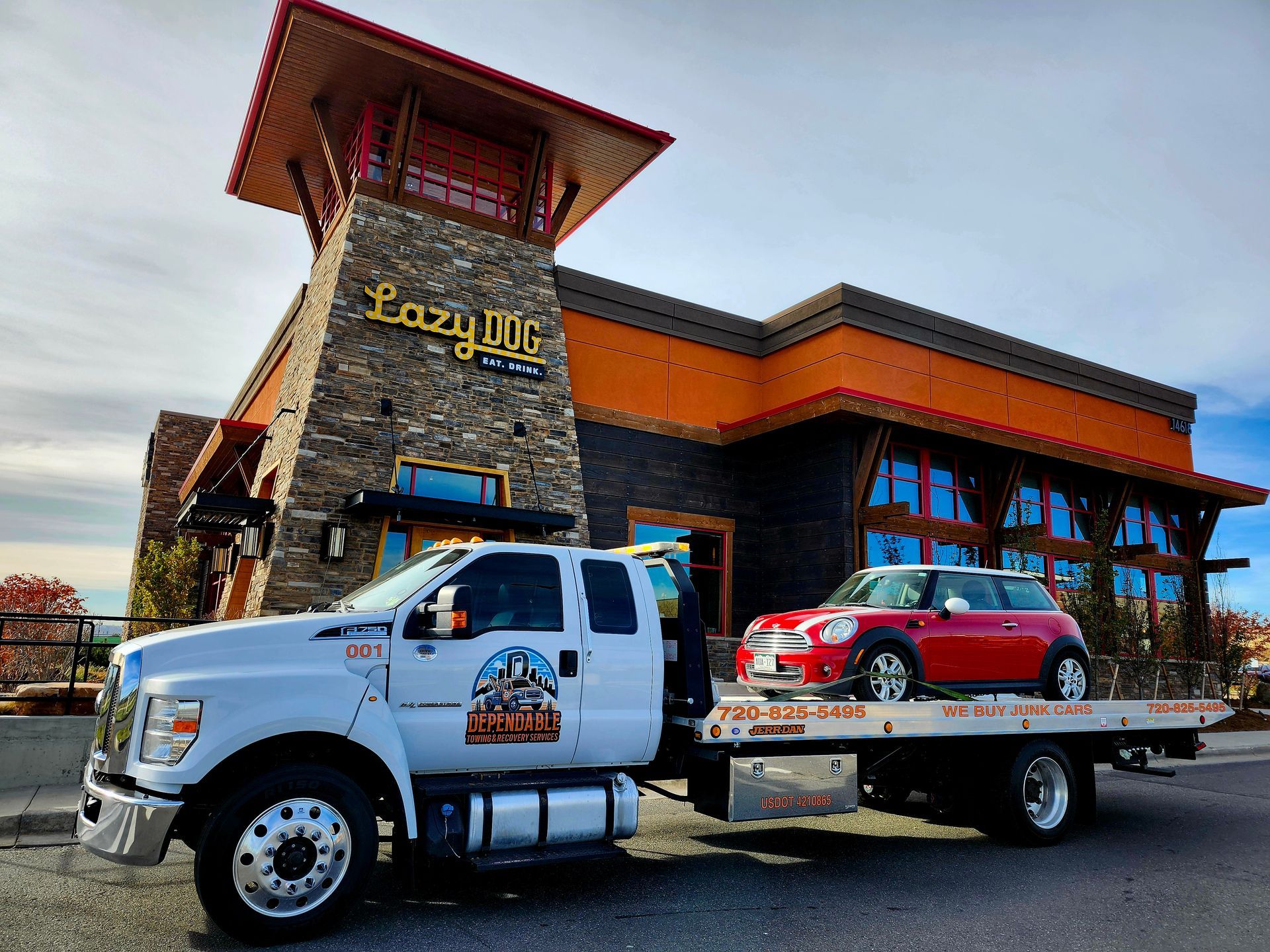 A flatbed tow truck carries a red Mini Cooper in front of a Lazy Dog restaurant with stone and orange trim.