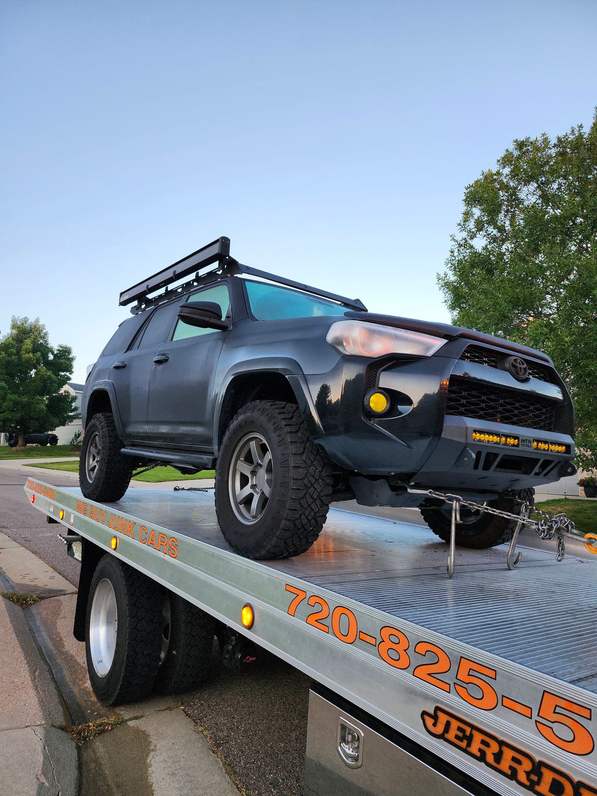 A black, modified Toyota 4Runner secured to the back of a flatbed tow truck on a suburban street.