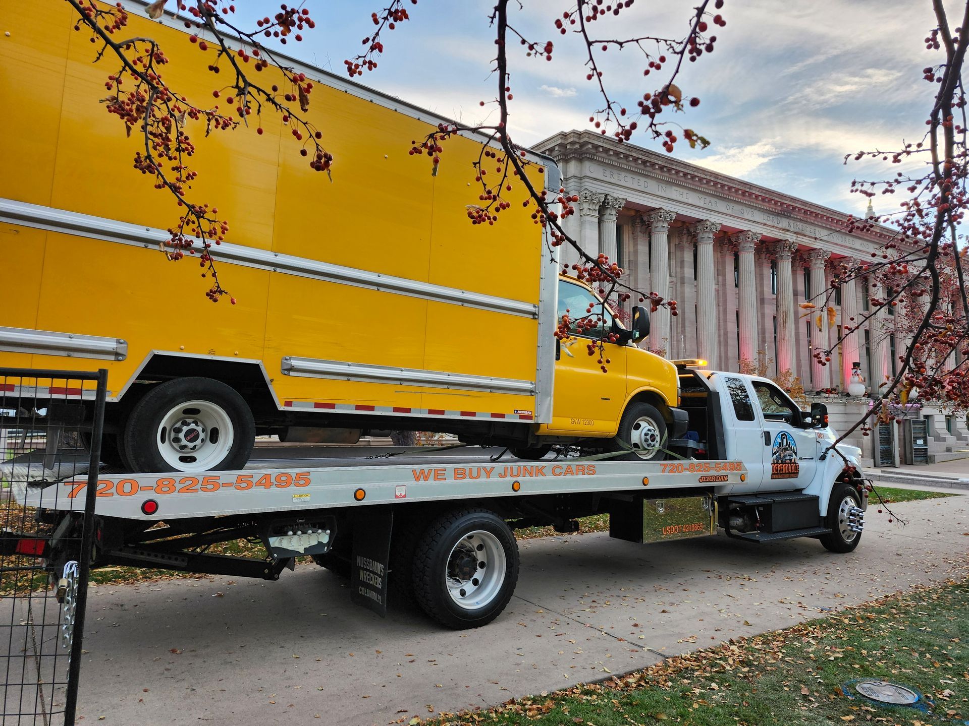 A yellow box truck being hauled on a flatbed tow truck in front of a grand stone building with columns.