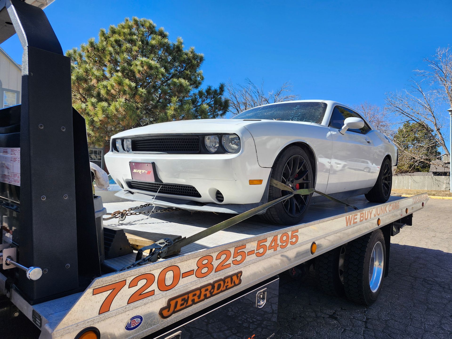 A white Dodge Challenger securely strapped onto the flatbed of a Jerr-Dan tow truck on a sunny day.