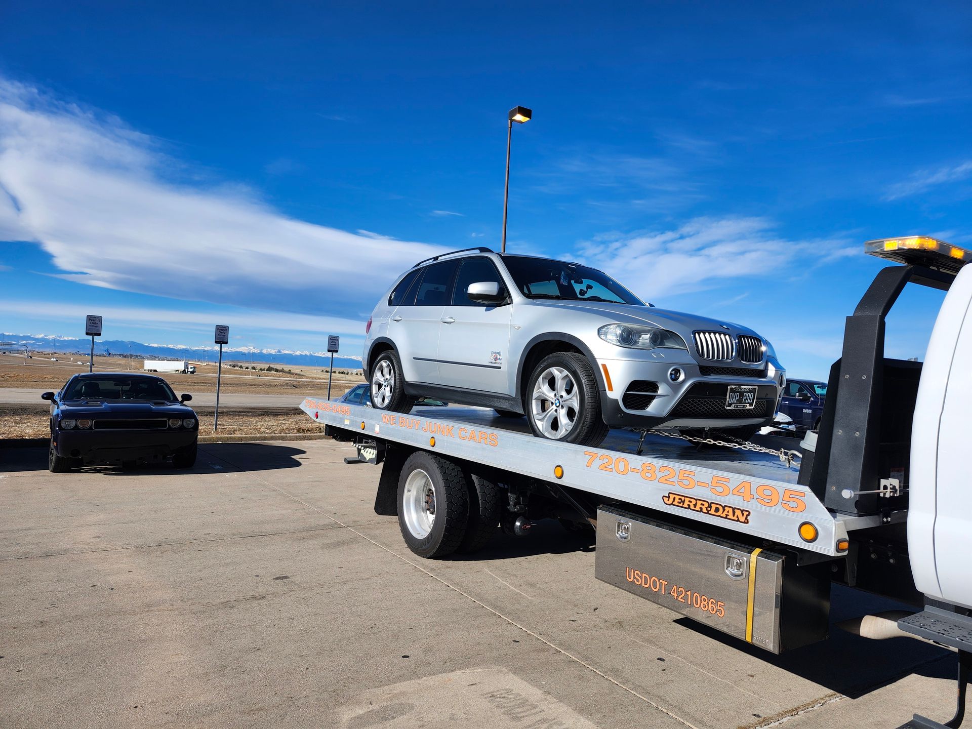 A silver BMW SUV loaded onto the flatbed of a tow truck in an outdoor parking lot under a bright blue sky.