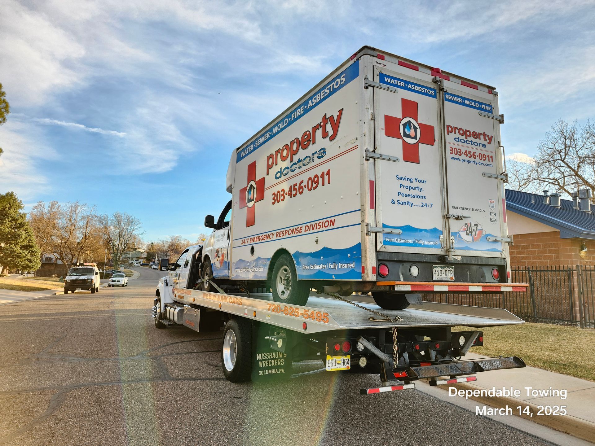 A white Property Doctors box truck is being towed on a flatbed tow truck on a suburban street.