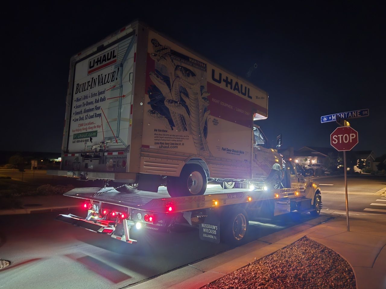 A U-Haul truck loaded onto a flatbed tow truck at night, parked near a stop sign on a residential street.