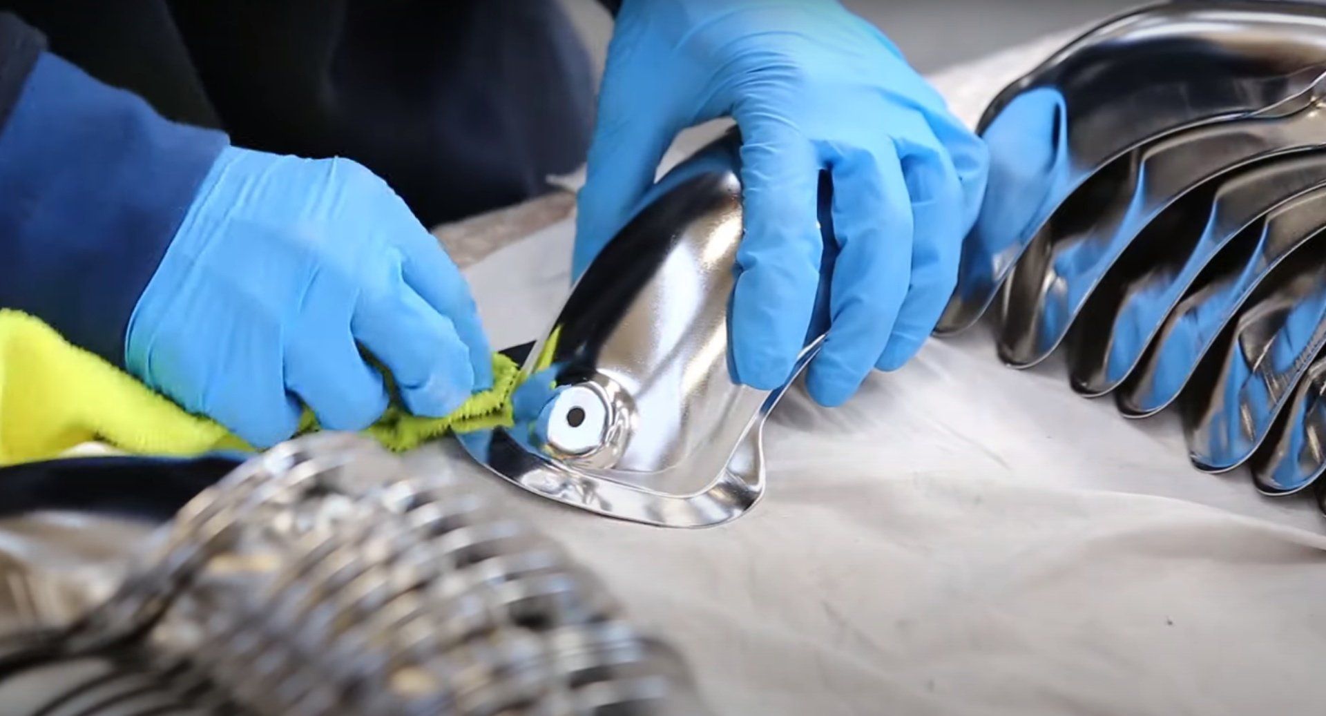 Mani con guanti blu che lucidano un oggetto cromato e lucente, probabilmente parte di un pezzo più grande.