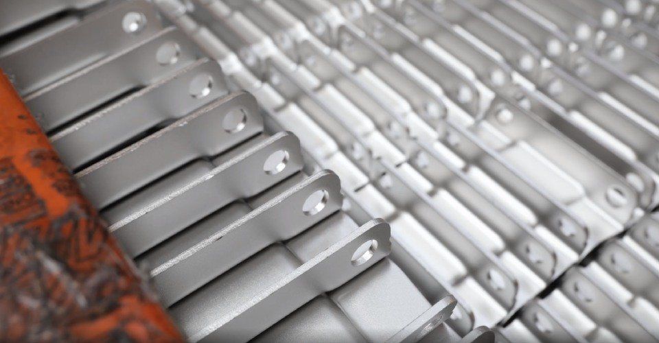 File di staffe metalliche argentate con fori, impilate su una superficie, probabilmente in una configurazione di fabbrica.